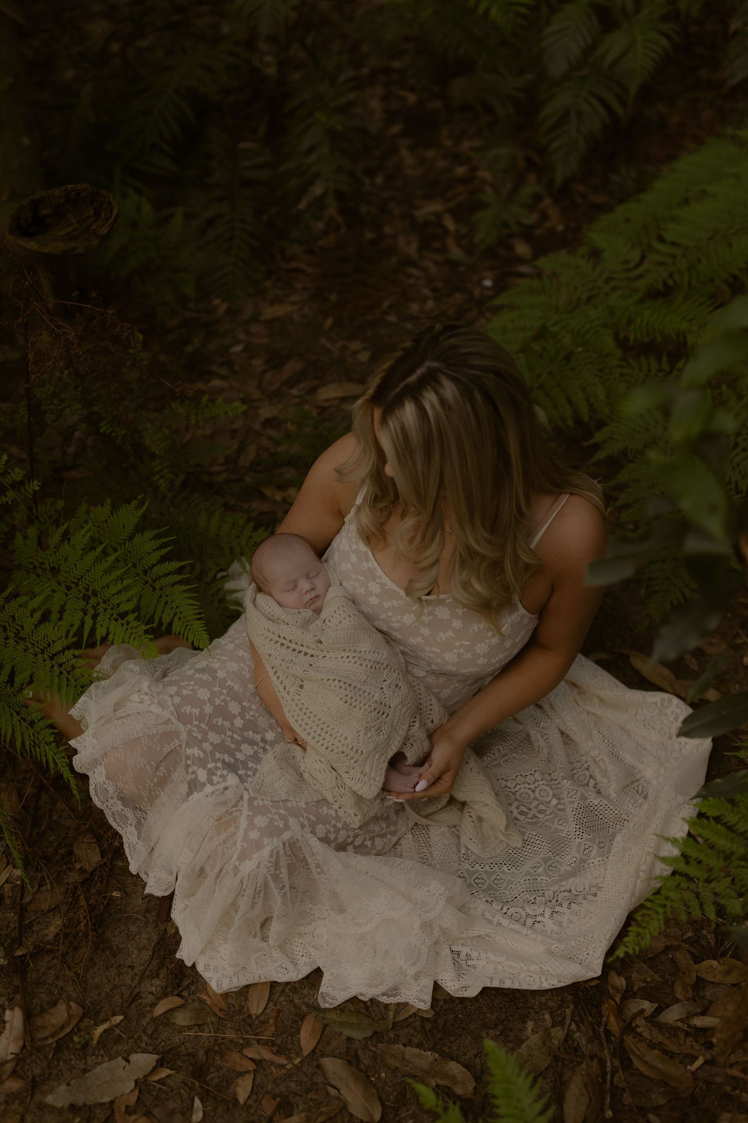 A mother in a lace dress cradles her newborn baby surrounded by lush ferns in a forest setting.