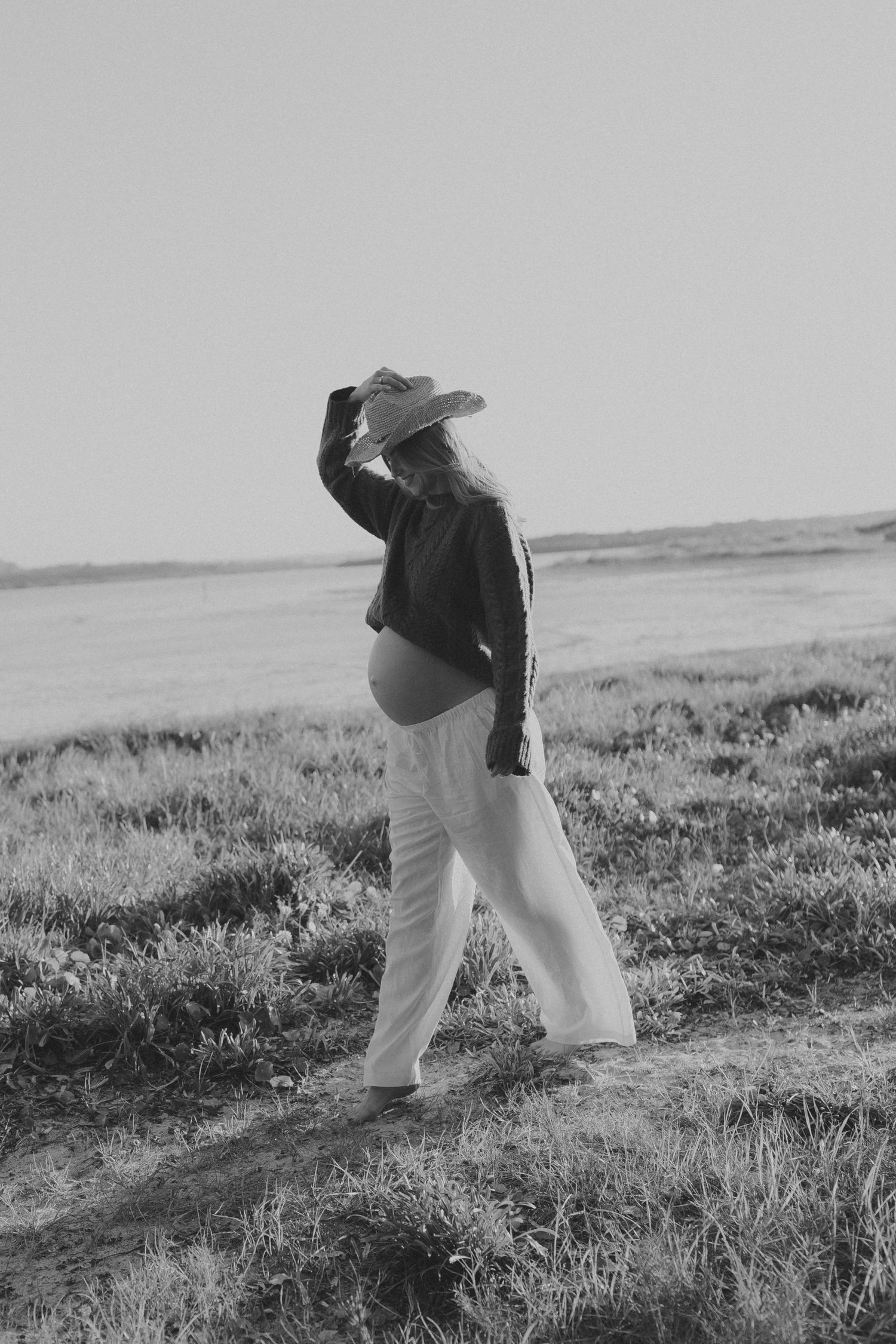 Black and white photo of a pregnant woman in a wide-brim hat and white pants standing in a field.
