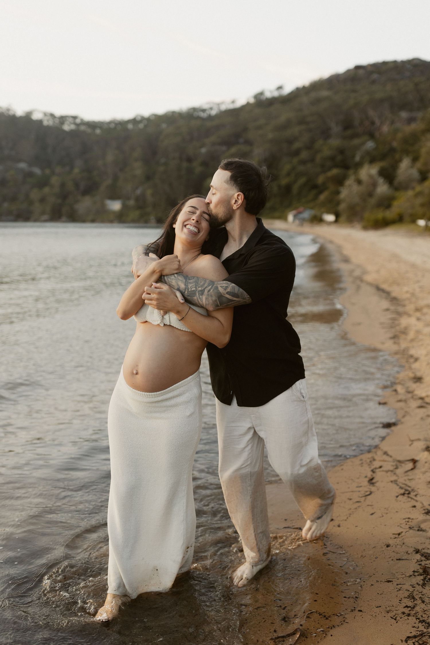 Maternity couple embraces joyfully on sandy beach with mountains and lake in background during golden hour.