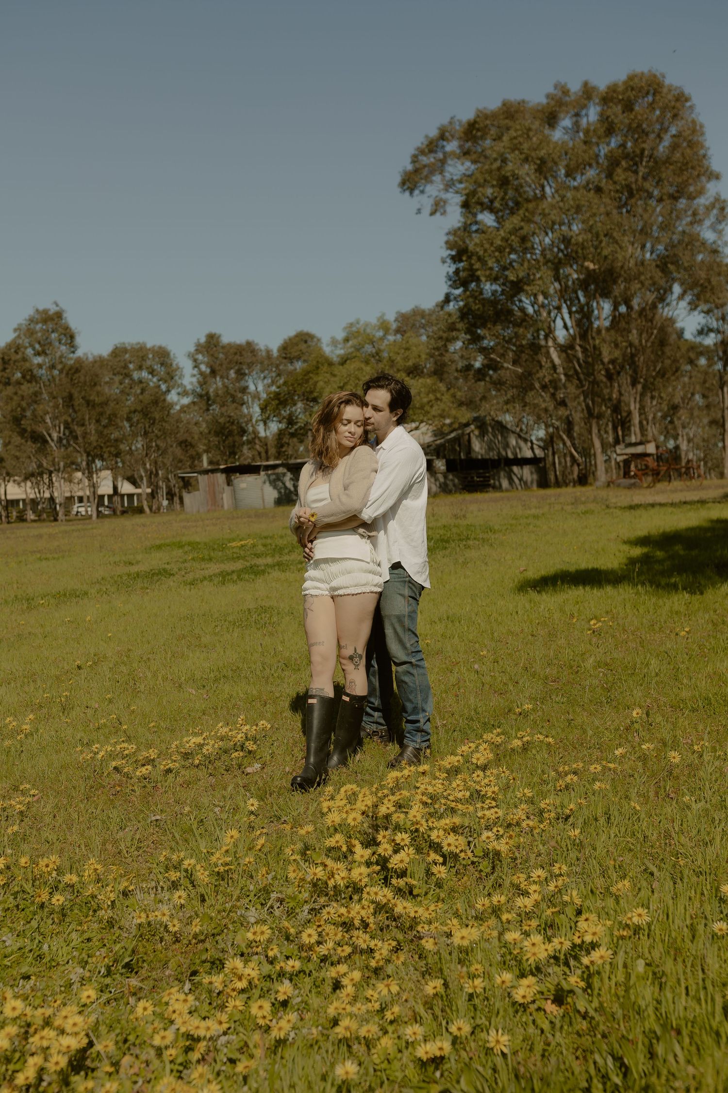 A couple embraces in a sunny meadow filled with yellow wildflowers and trees in the background.