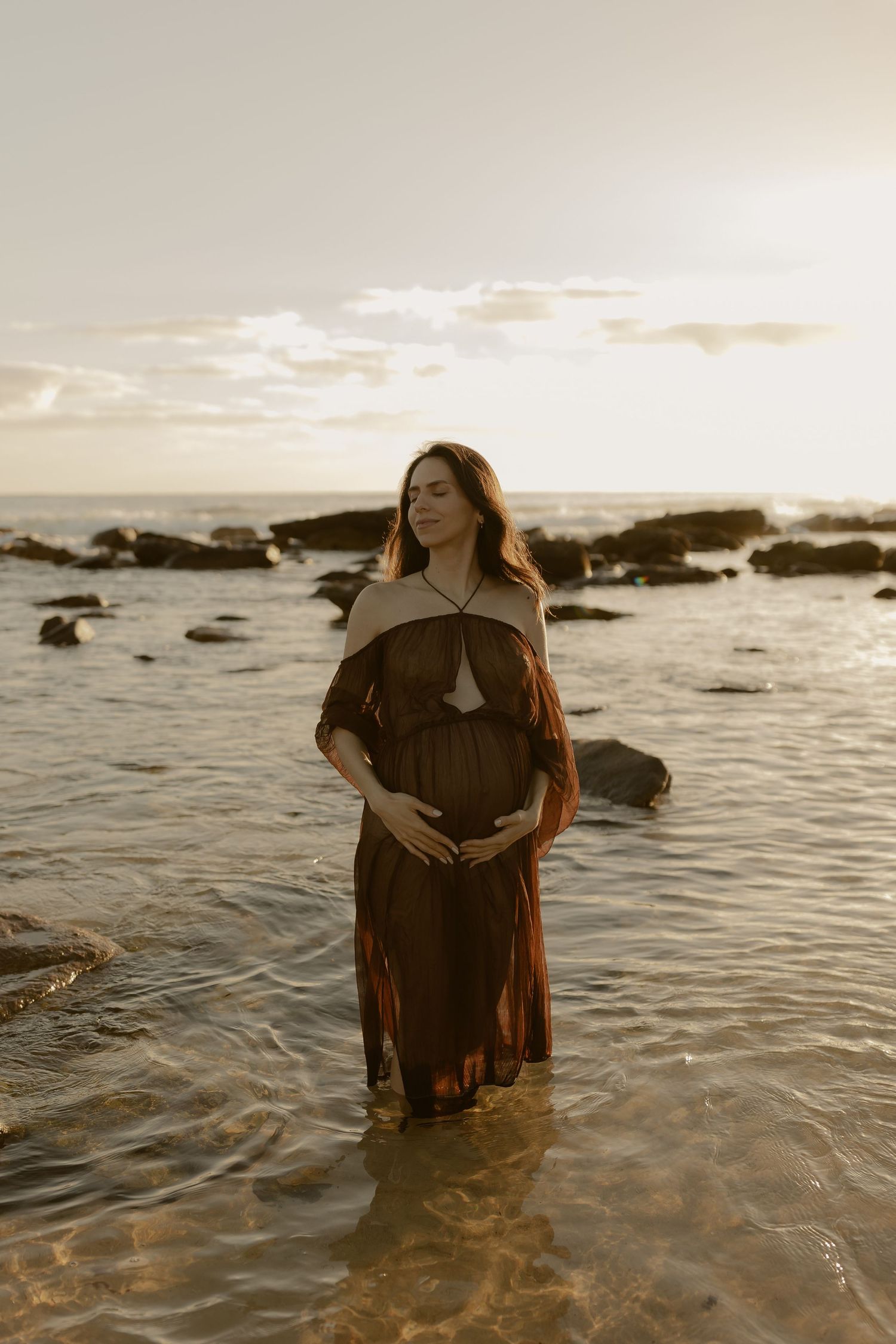 A person stands in shallow ocean water wearing dark clothing during a sunset beach photoshoot.