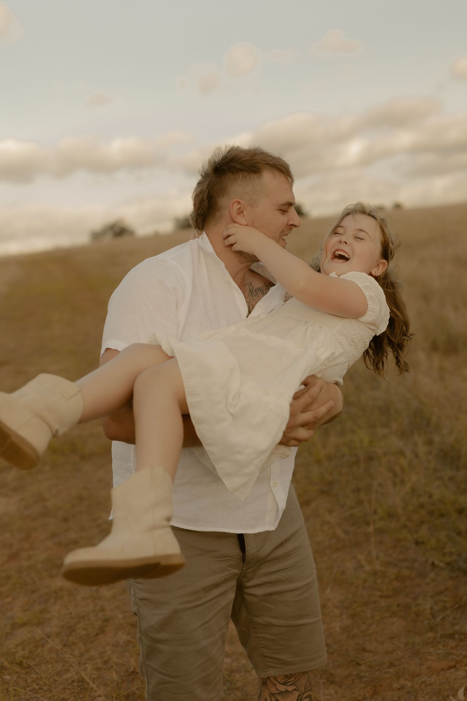 A joyful couple in white clothing share a playful moment together in a grassy field at sunset.