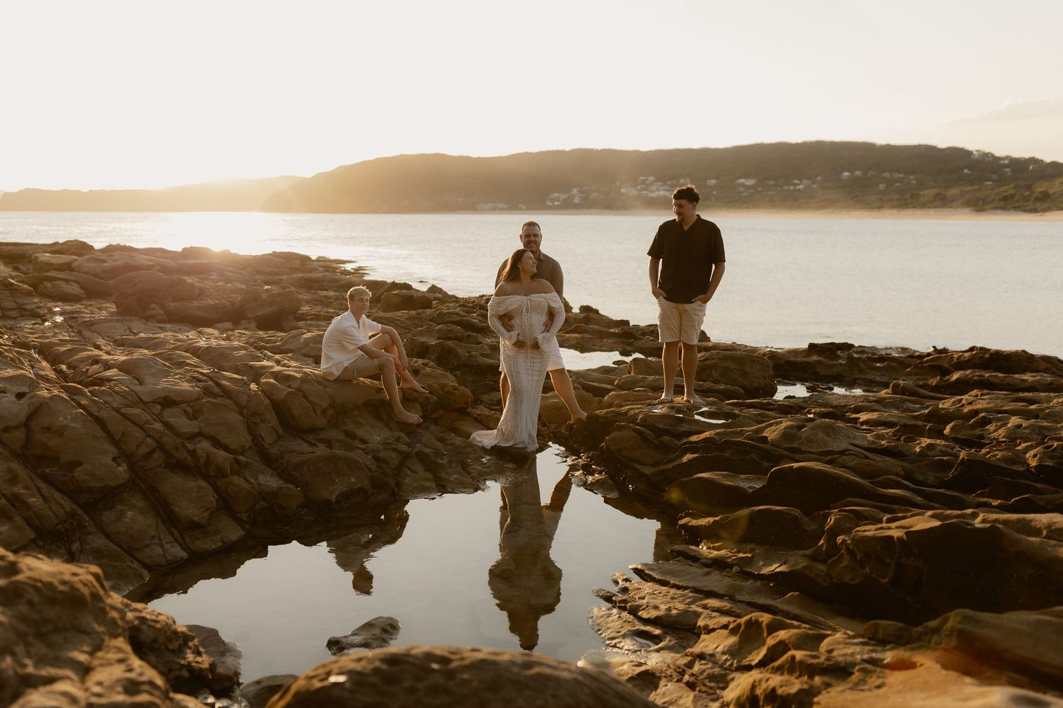 Two people stand on rocky coastal cliffs overlooking a serene ocean view at sunset.