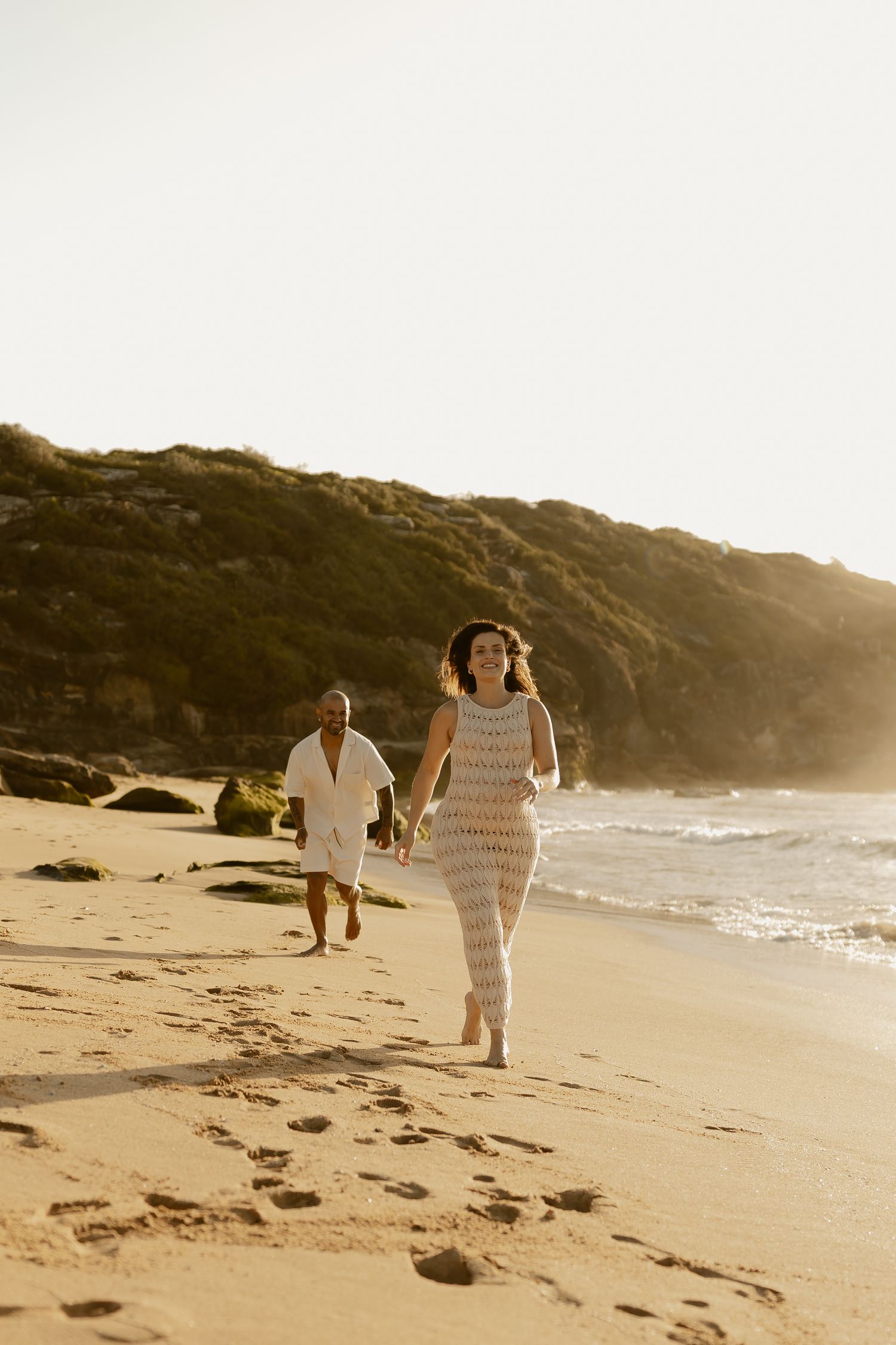 Couple walking along scenic beach at sunset with golden light and cliffs in background.