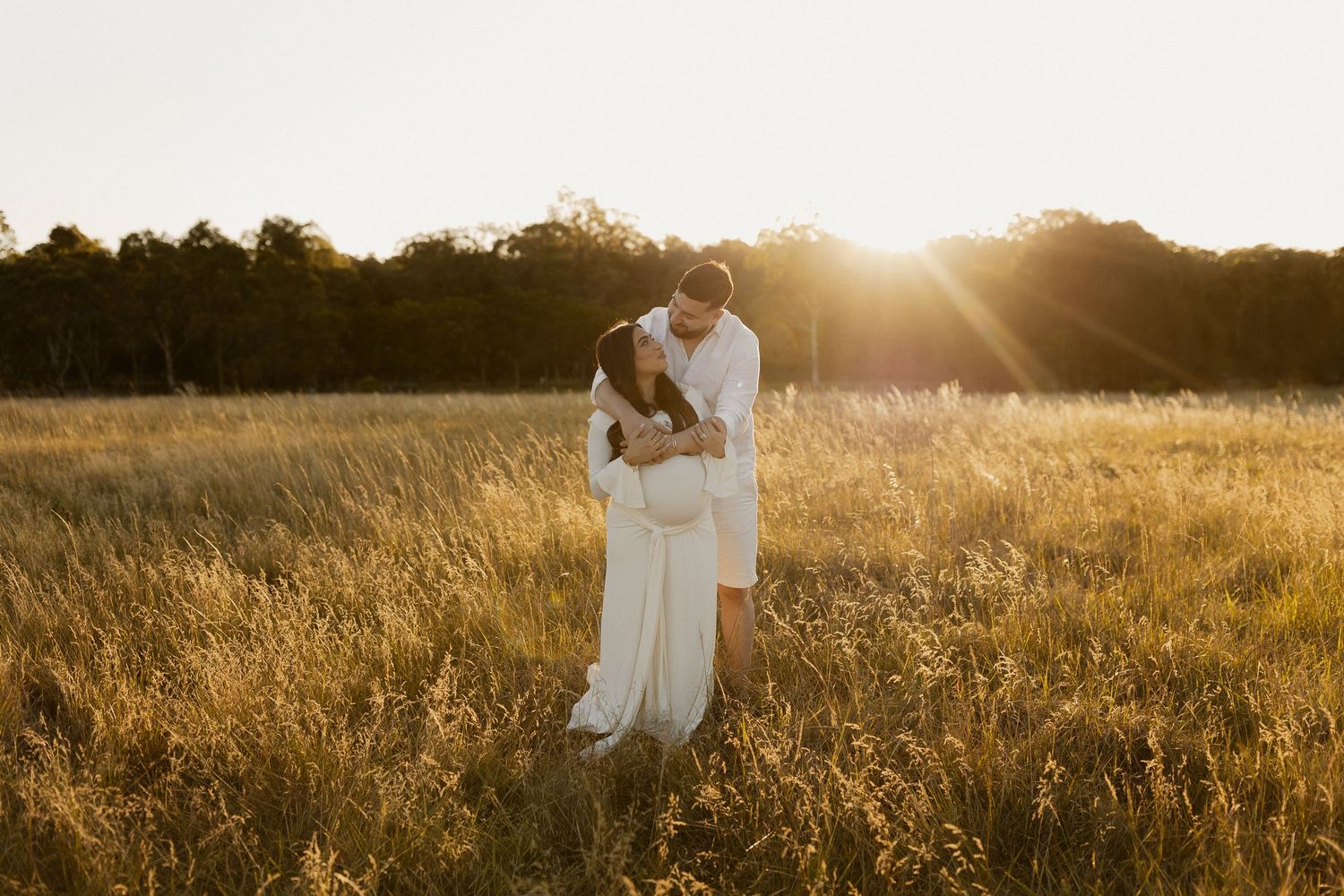 A romantic couple embraces in a golden sunlit field at sunset during a dreamy outdoor photoshoot.