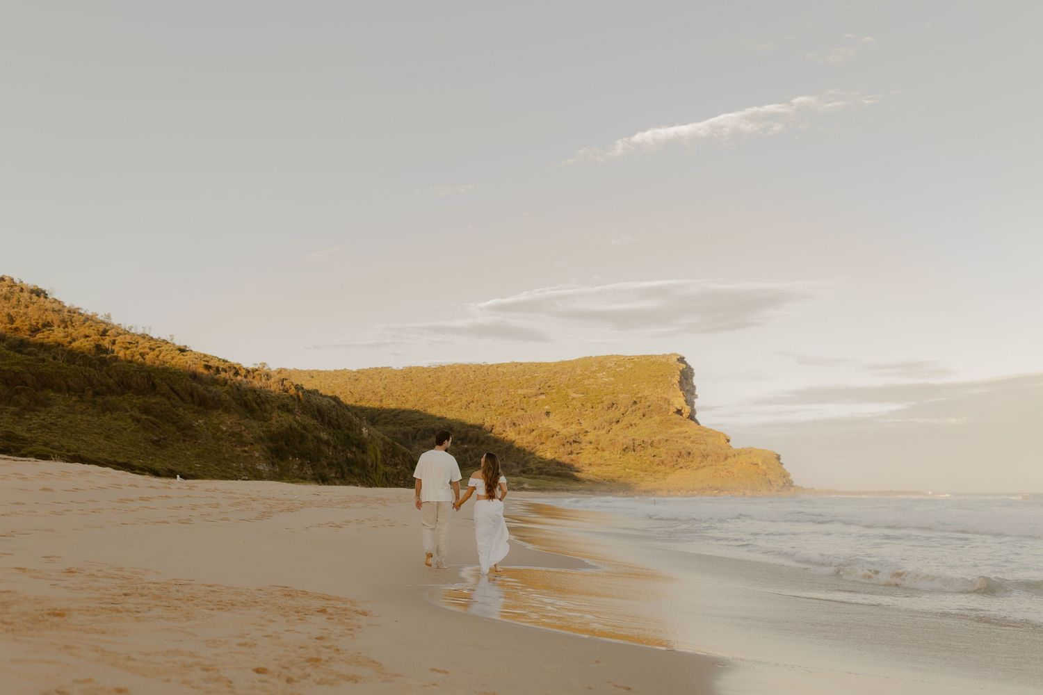 A couple in white clothing walks along a scenic beach at sunset with rolling hills in the background.