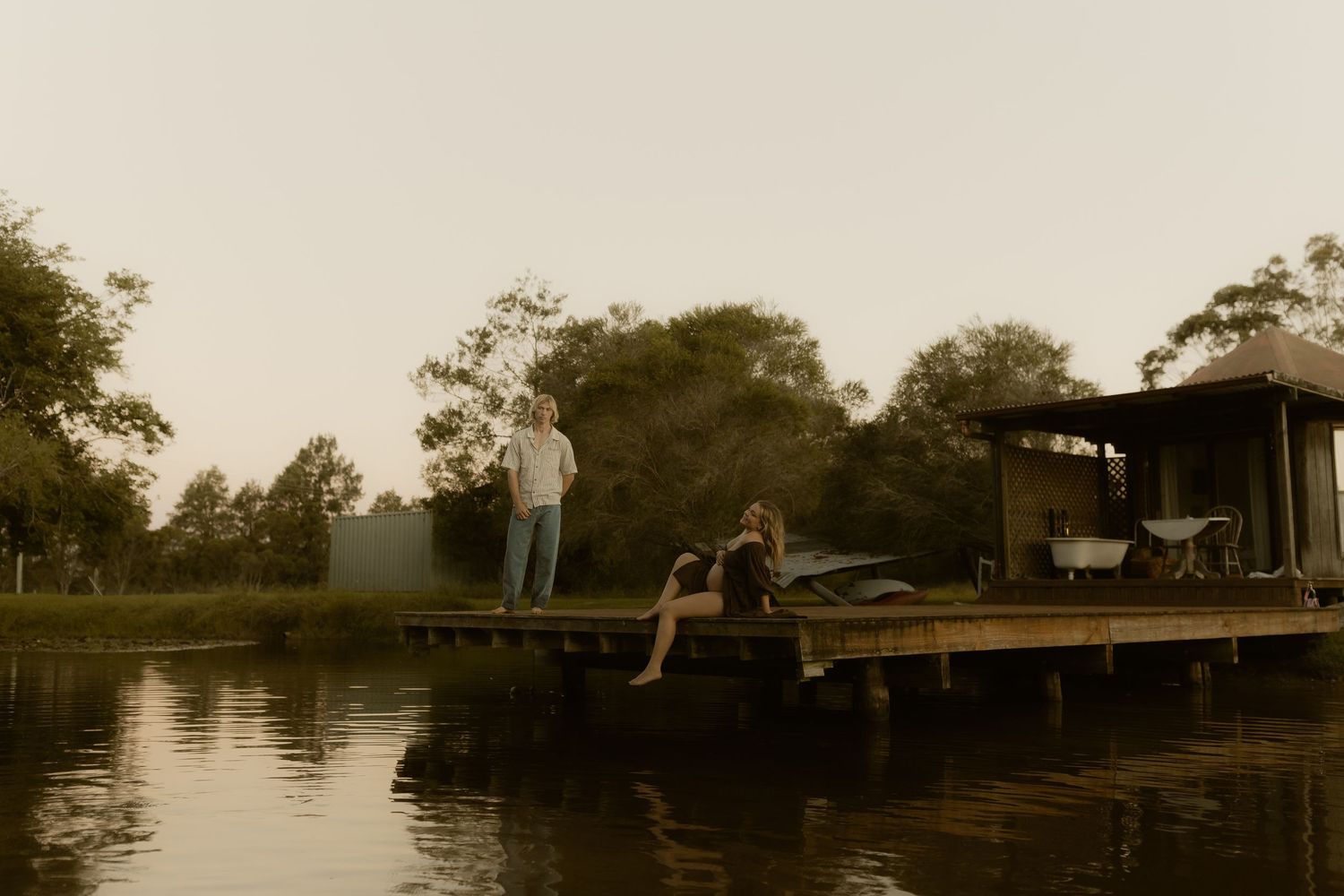 People relaxing on wooden dock by lake during sunset with rustic building in background.