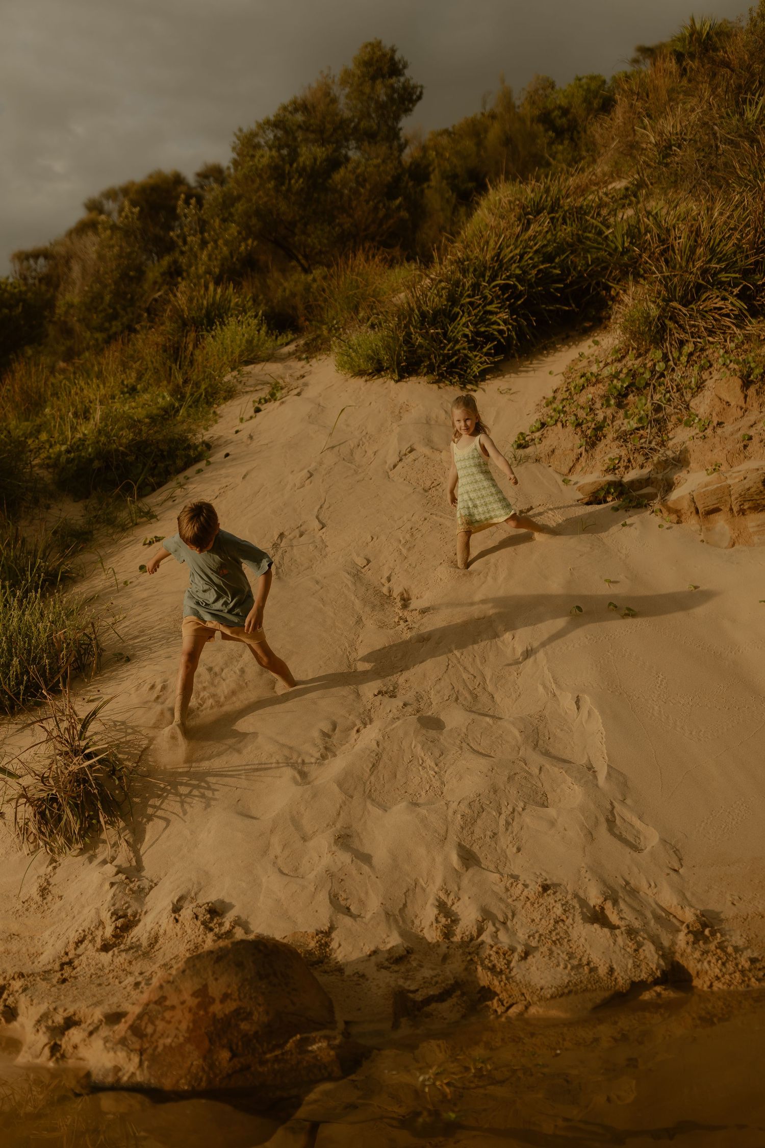 Children play on sandy slopes near coastal vegetation during sunset.