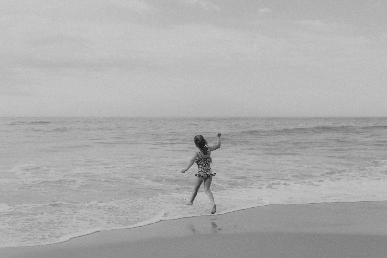 A silhouetted figure dances joyfully at the edge of ocean waves on a misty beach in black and white.