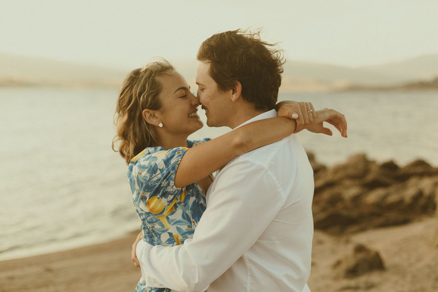 A romantic couple embraces at sunset on a rocky beach with golden light illuminating the scene.