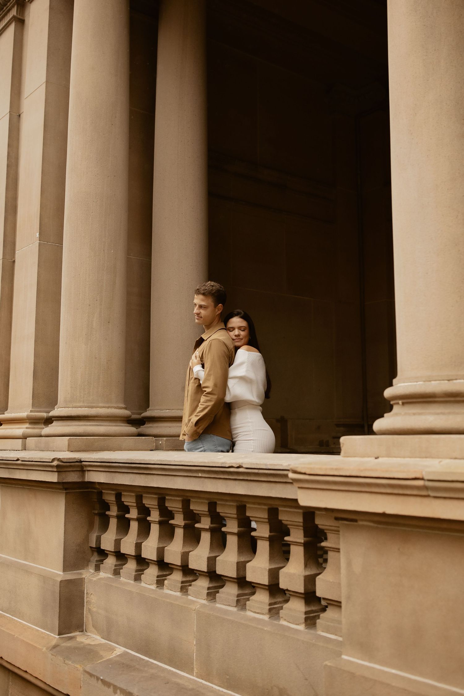 A couple poses against classic stone columns and balustrade in vintage-style photography.