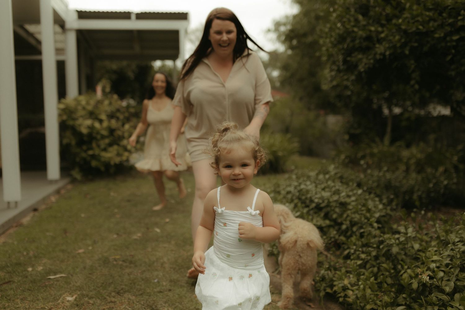 A toddler in a white dress walks through a garden path on a sunny afternoon.