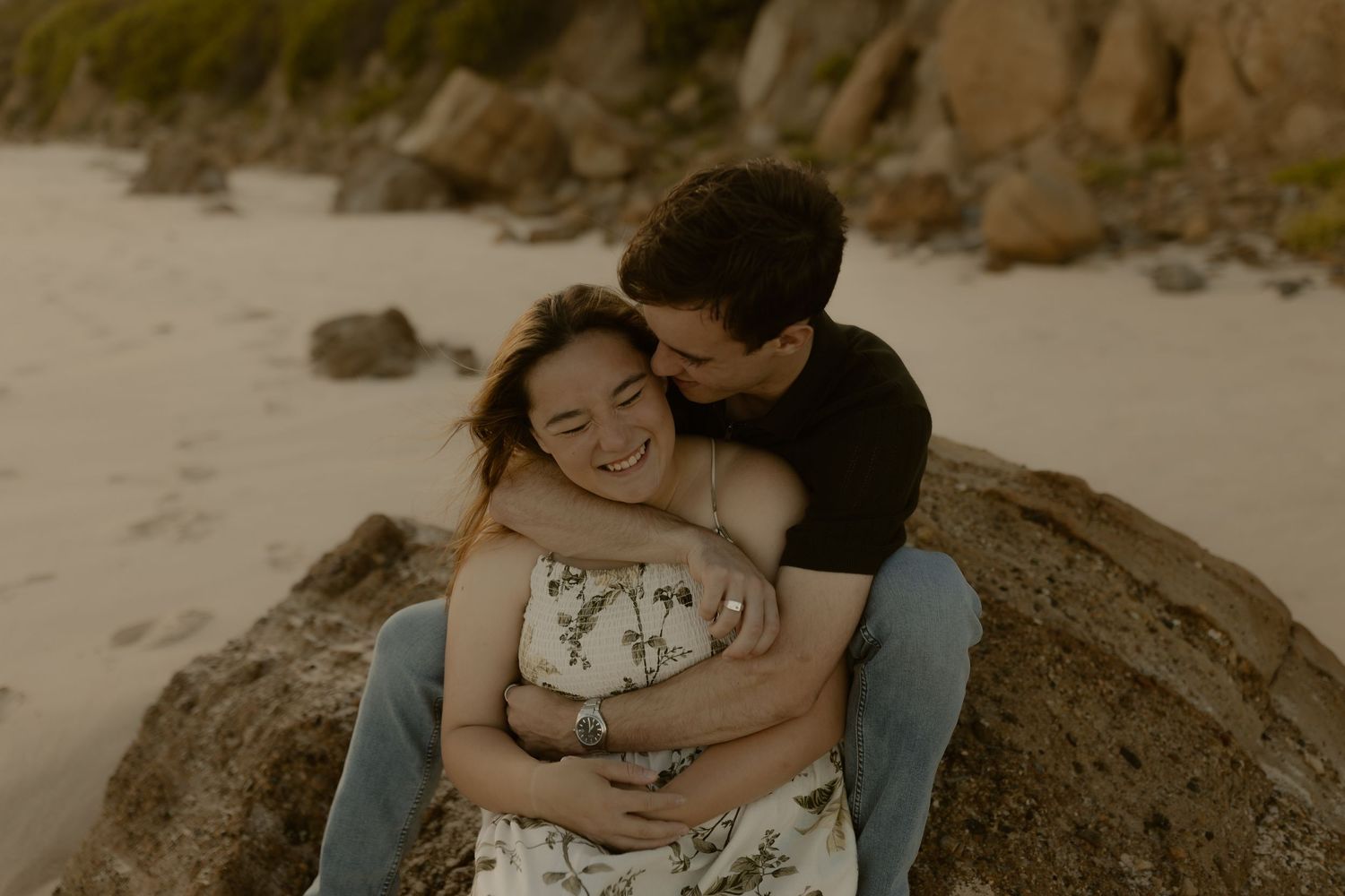A couple embraces on a rocky beach during a romantic sunset photoshoot in sepia tones.