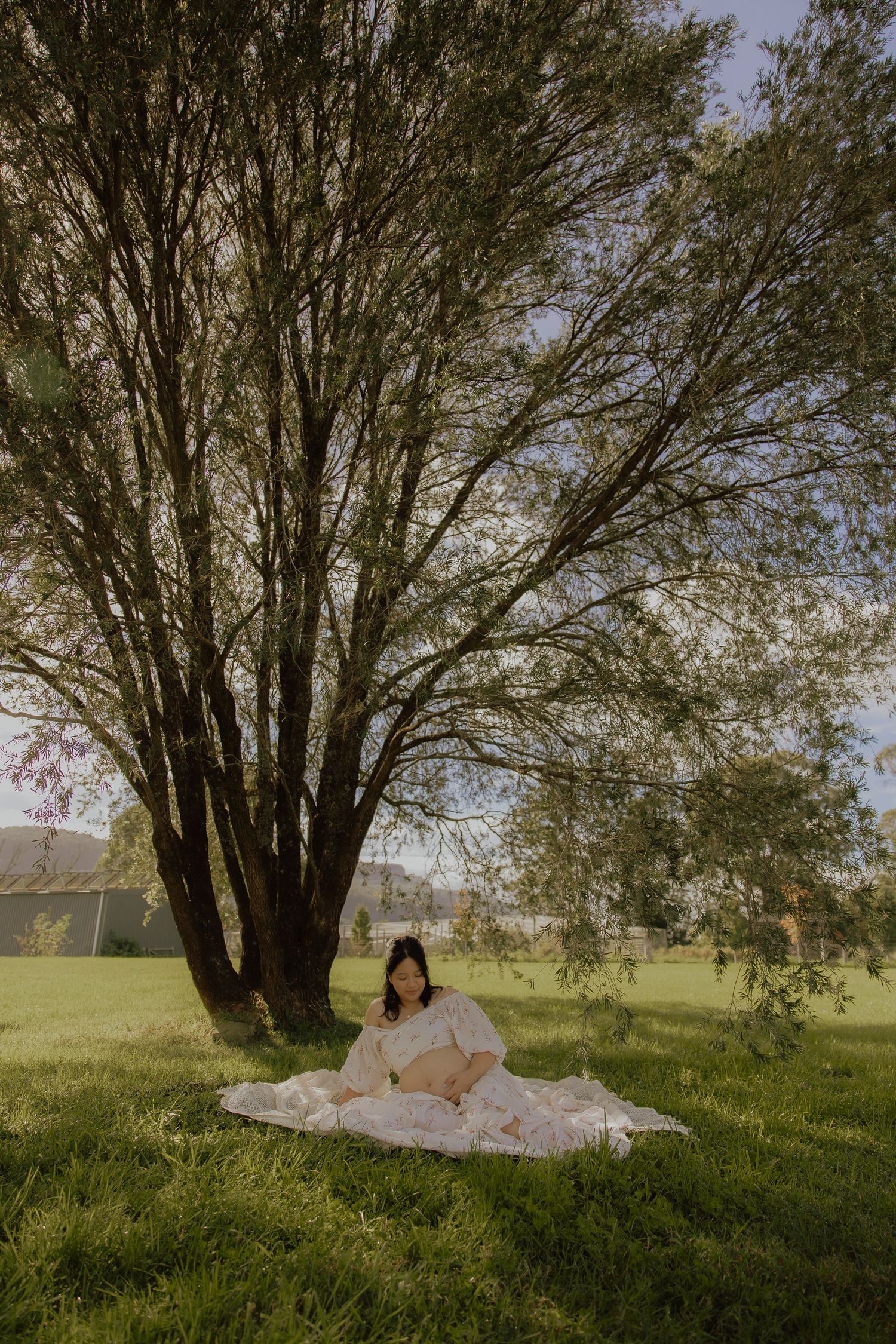 Pregnant woman in white dress sitting on blanket under large tree in sunny green park.