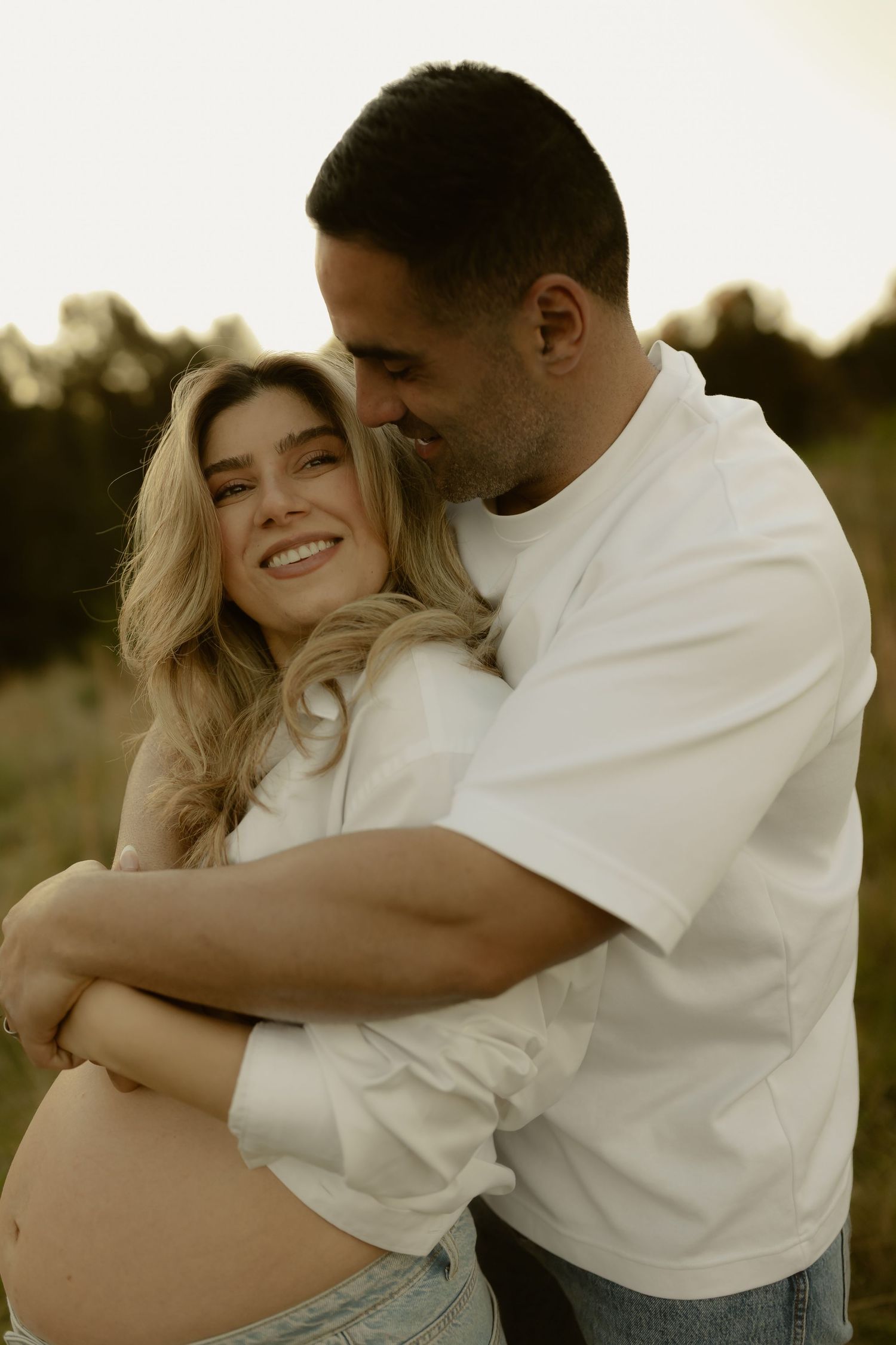 Couple embracing outdoors at golden hour, pregnant woman smiling as partner holds her from behind.