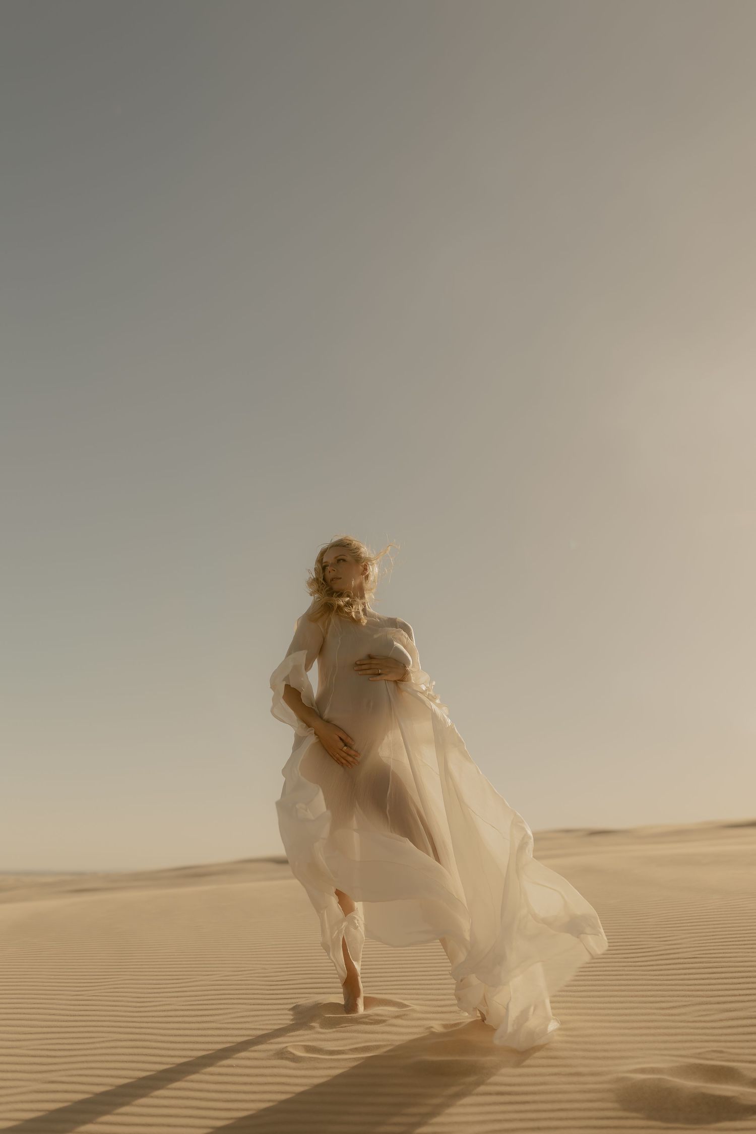 Pregnant woman in flowing white dress walking gracefully across golden sand dunes in a serene desert landscape.