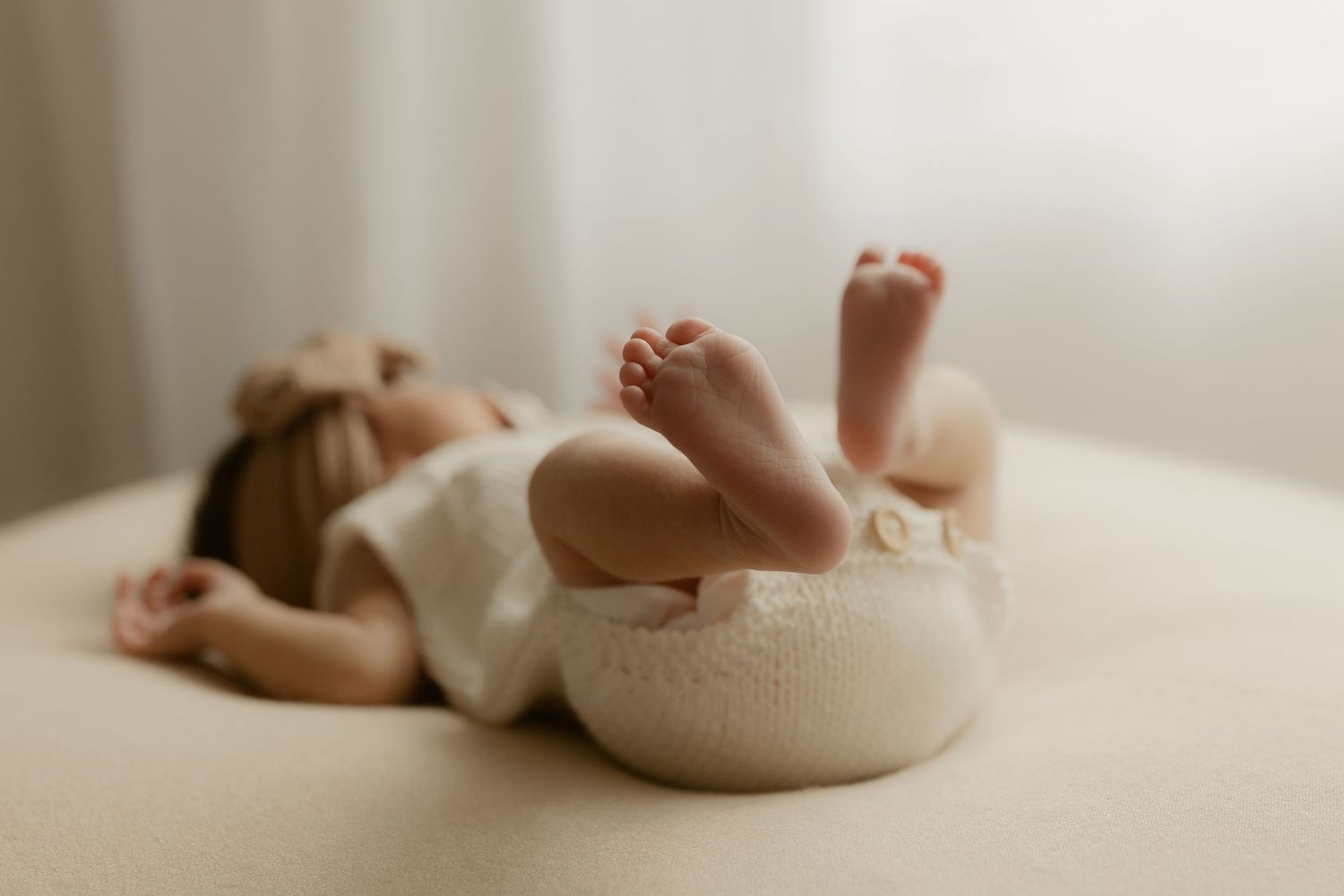 Newborn baby sleeping peacefully in a cream knit bowl, tiny feet raised, soft natural light photography.