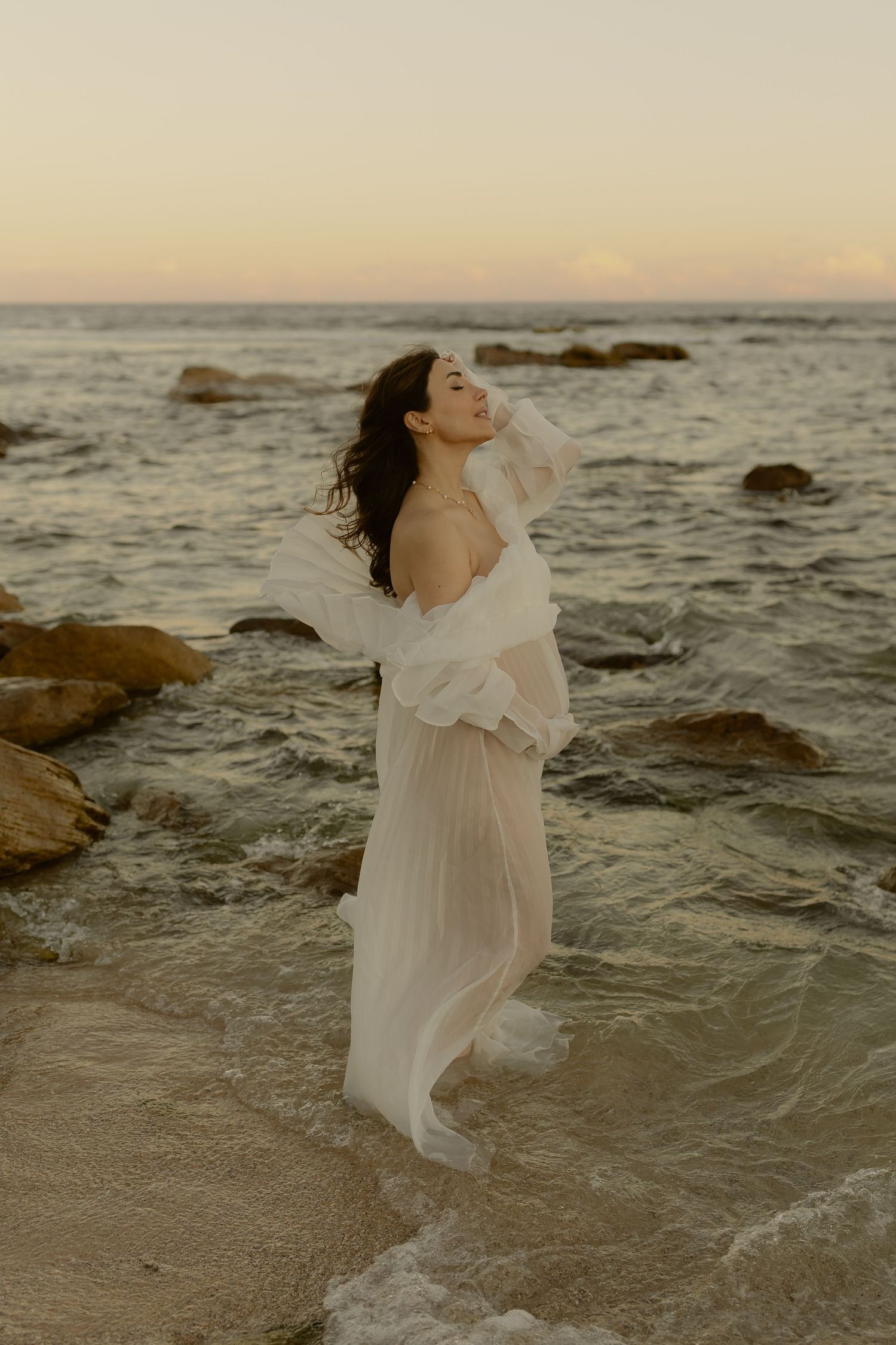 Woman in flowing white dress standing in ocean waves at sunset with rocks in background.