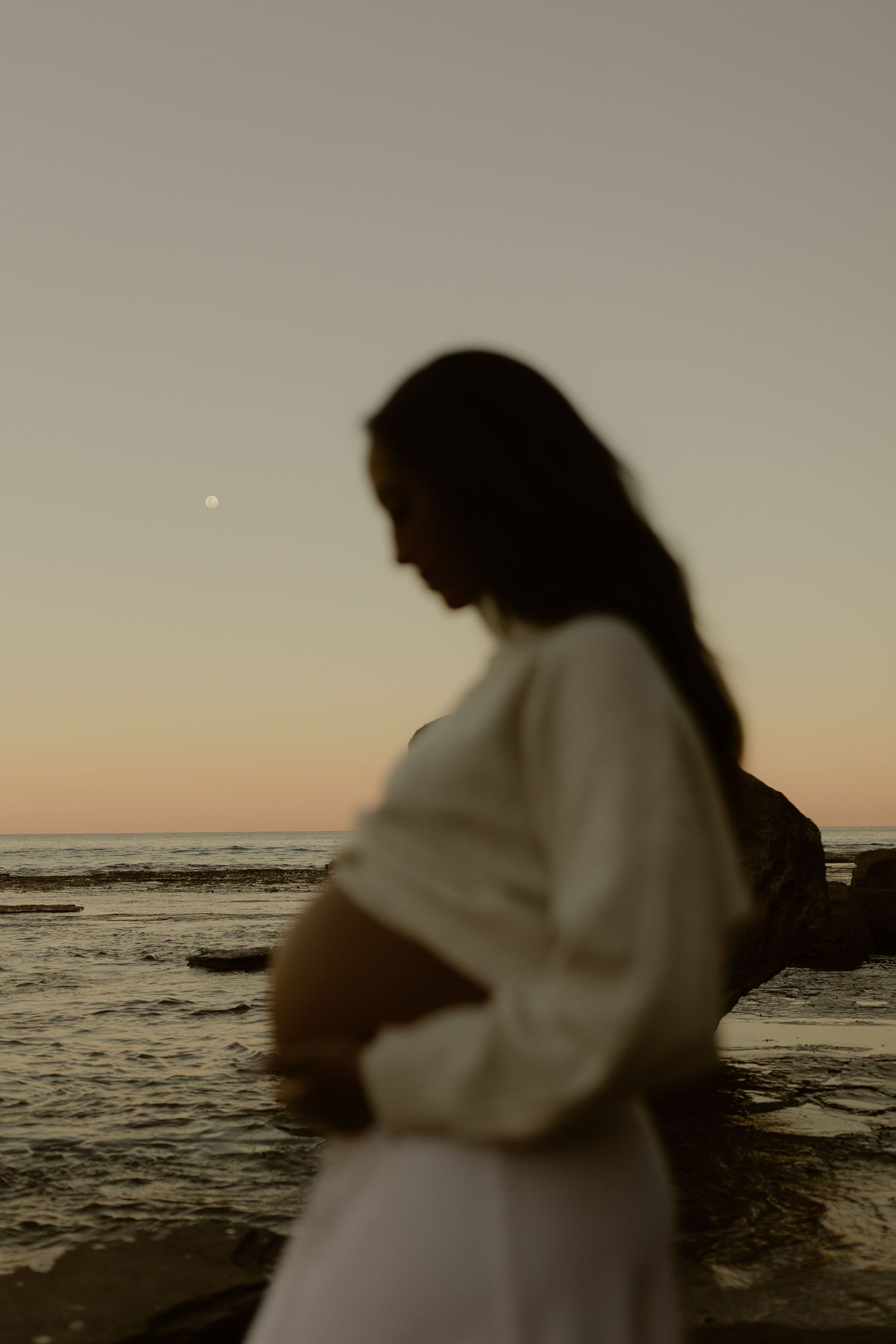 Pregnant woman in white dress cradles bump at ocean sunset with moon visible.