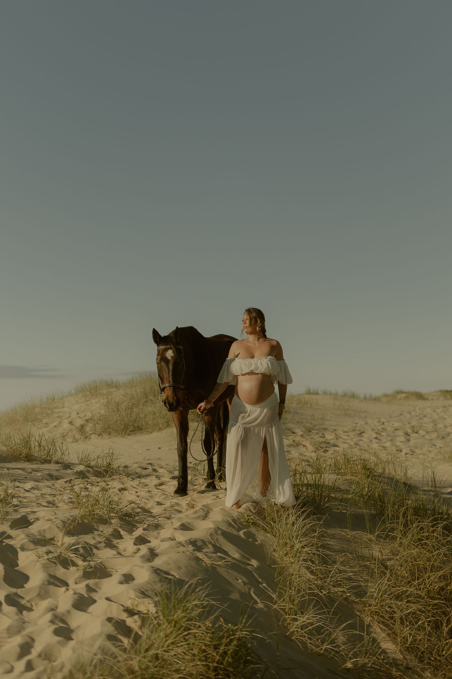 Pregnant woman in white dress walks with brown horse on sandy dunes under a clear sky.