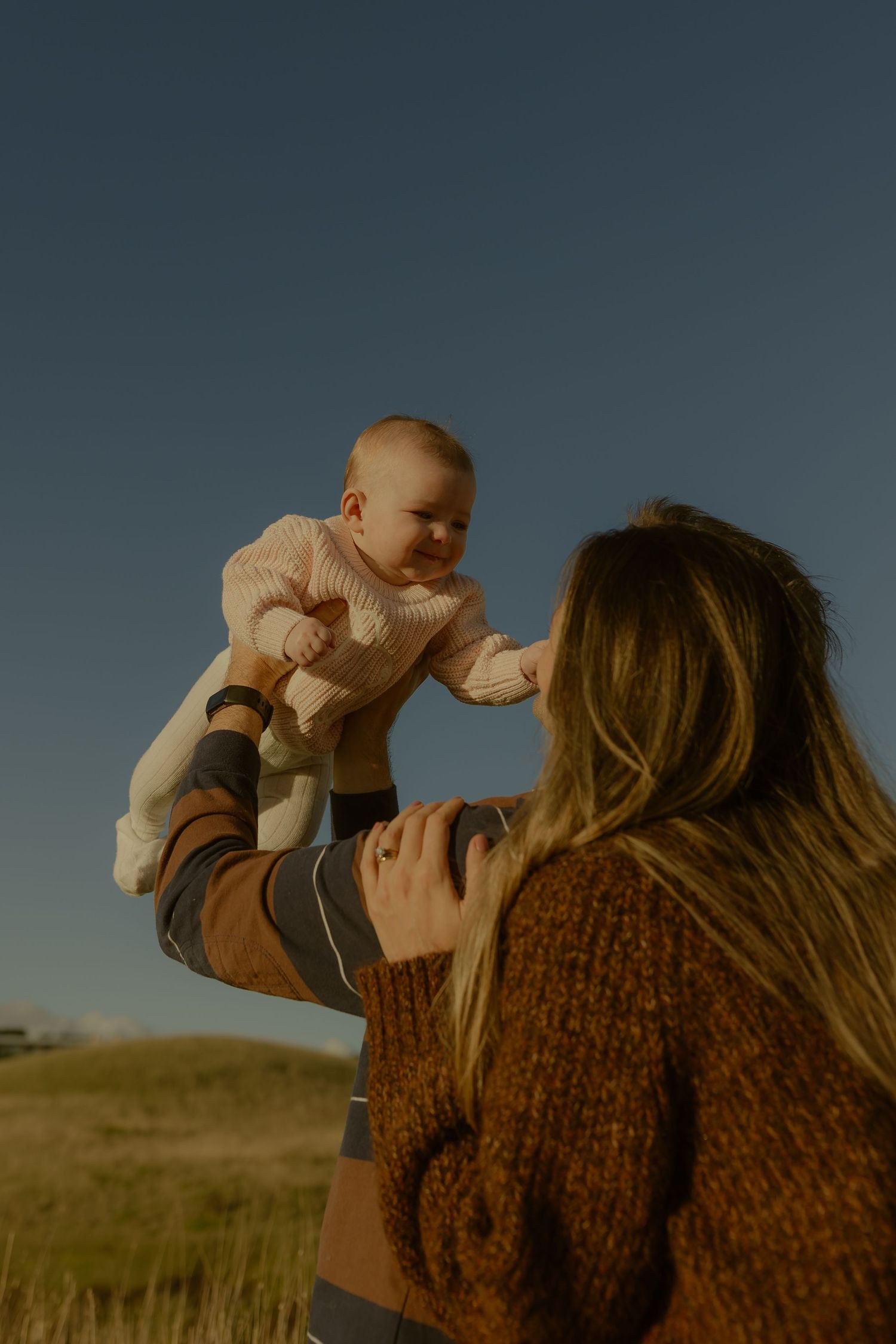 A mother lifts her smiling baby in the air outdoors at sunset in a grassy field.