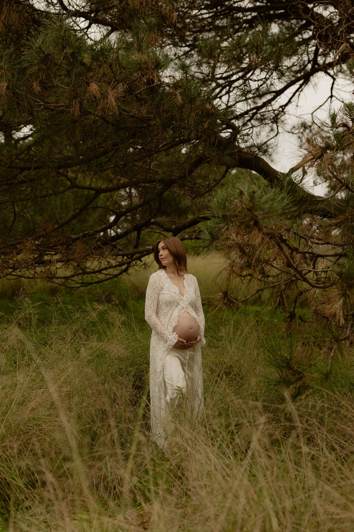 Pregnant woman in white lace dress standing among tall grass and pine trees.