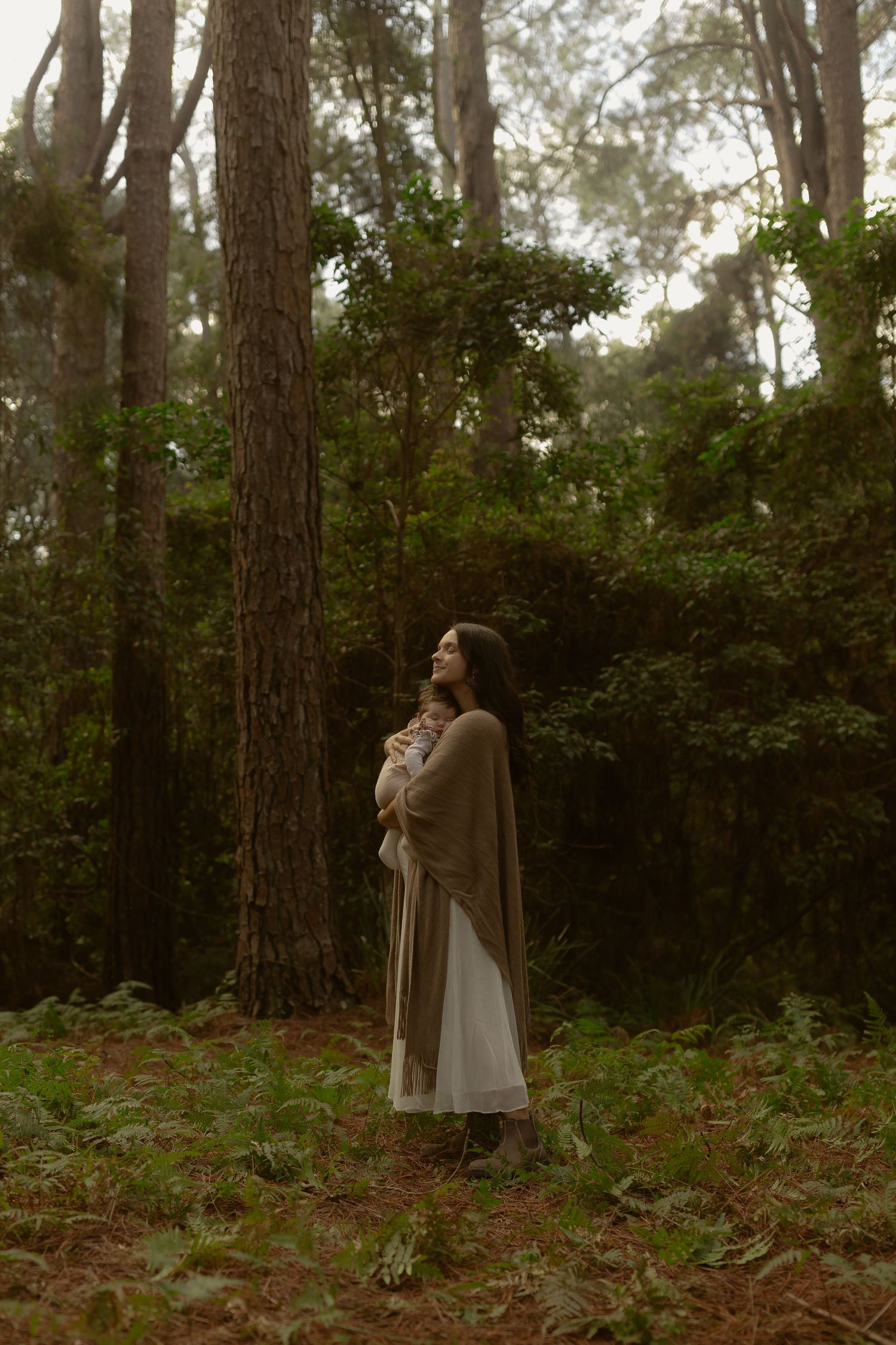 Woman in white dress and tan wrap holds newborn baby while standing in forest.