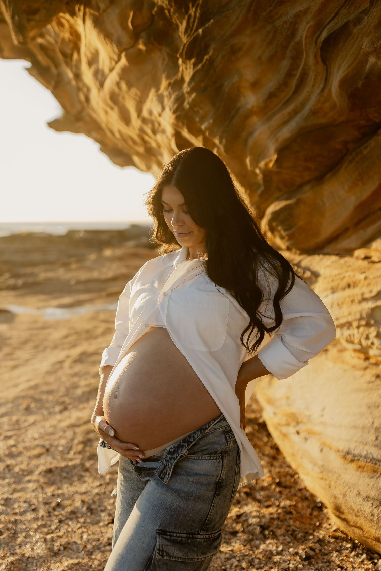 Pregnant woman in white shirt and jeans cradles her baby bump against golden sandstone rocks at sunset.