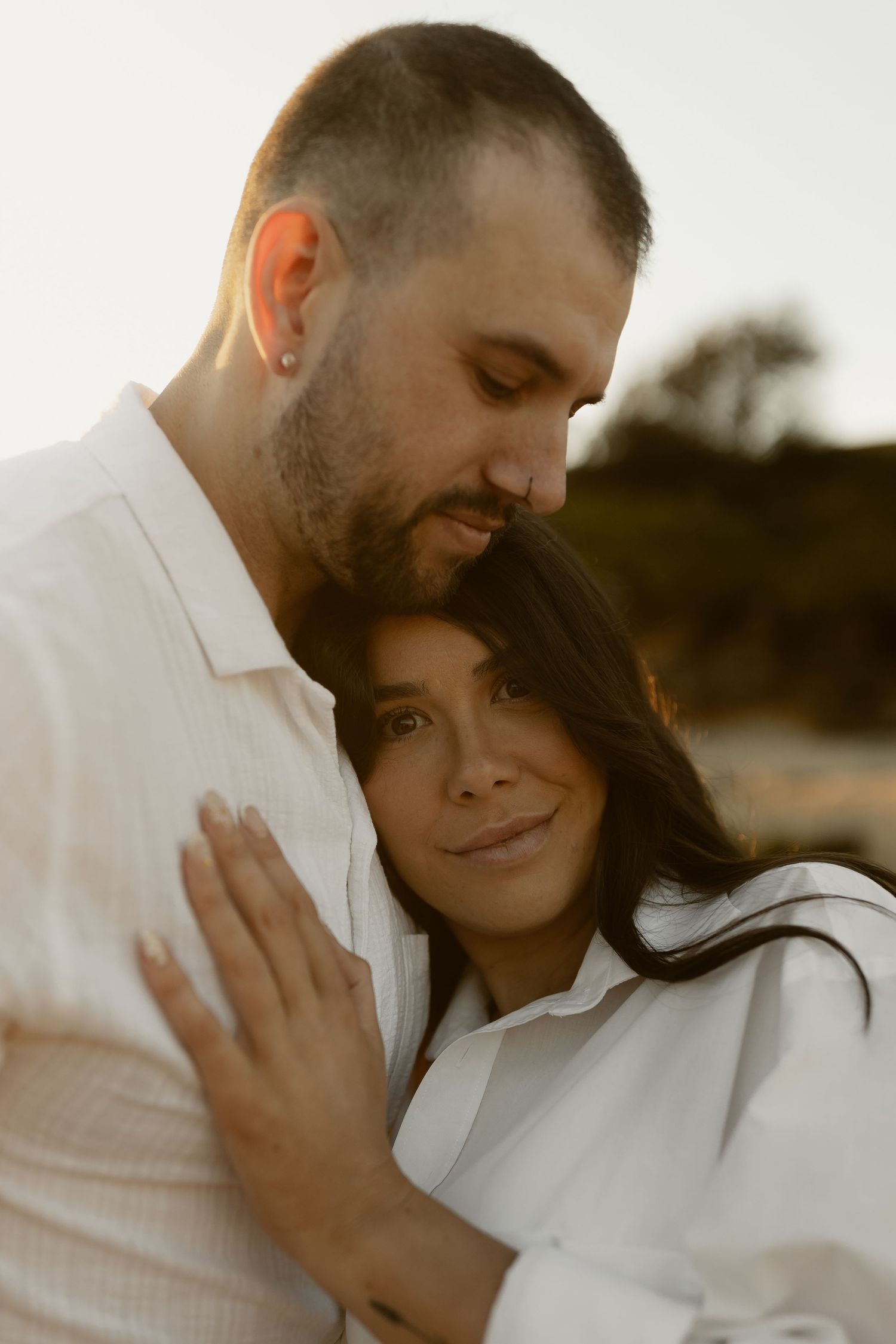 Couple embracing tenderly at sunset, woman in white shirt rests head on bearded mans chest outdoors.