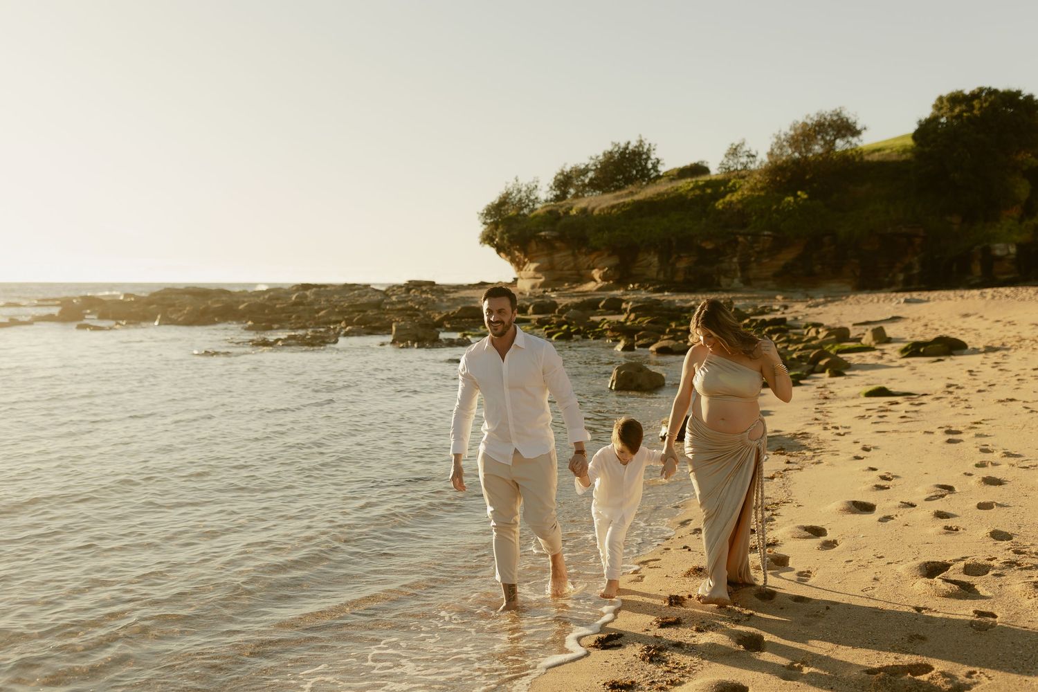 Family of three walking hand-in-hand along a sandy beach at golden hour, with rocky cliffs and calm ocean waves.