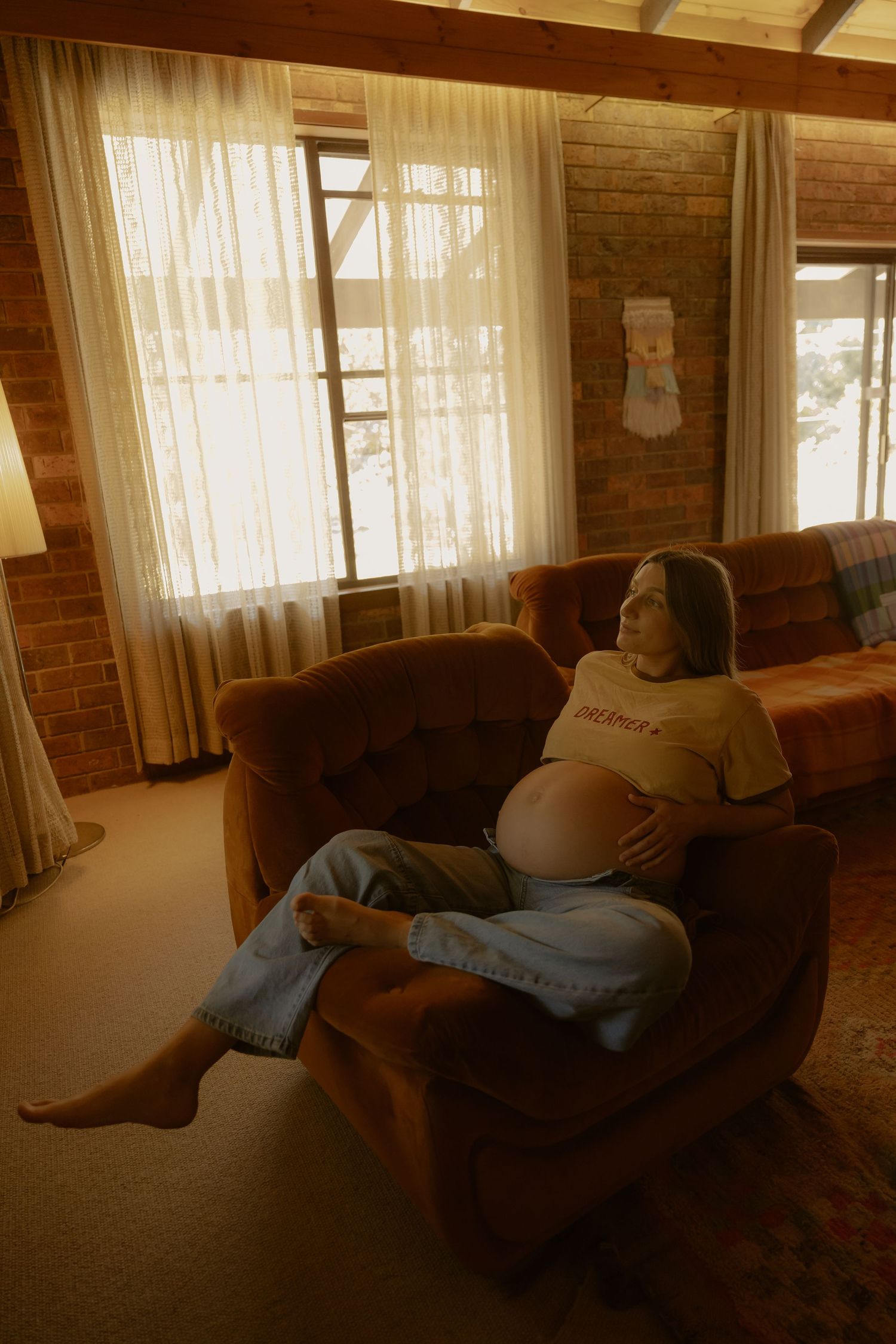 Pregnant woman relaxing in a brown armchair in a warm, rustic living room with brick walls and sheer curtains.
