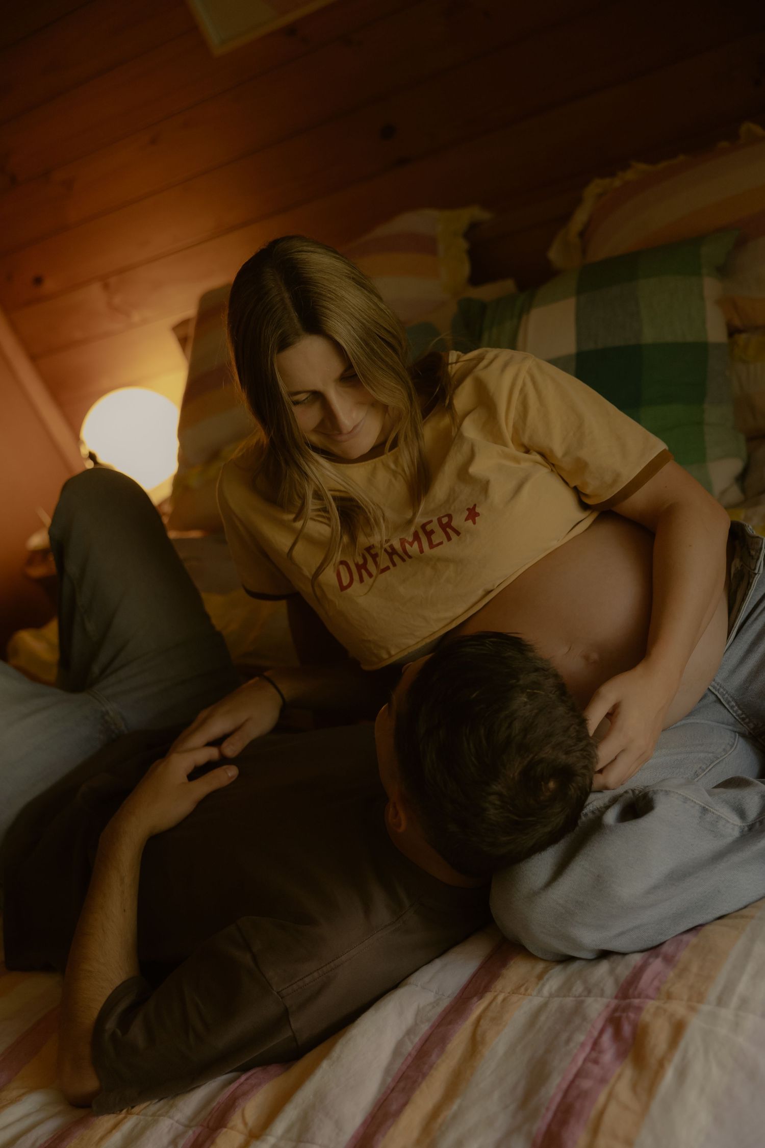 A couple relaxes on a bed in warm lamplight, woman in a yellow Dreamer shirt smiling down at her partner.