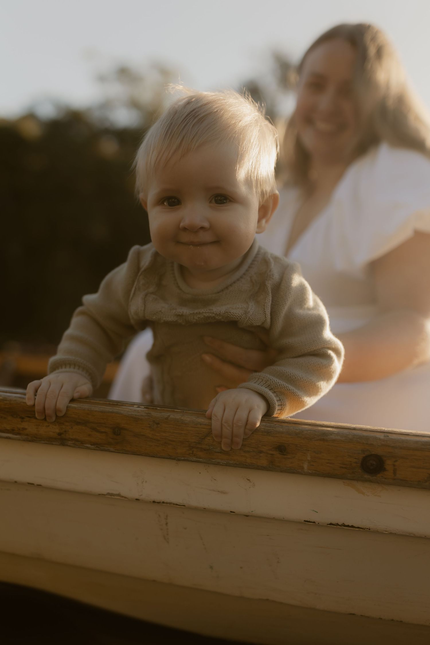 Smiling baby in a beige sweater standing at the edge of a wooden boat with a blonde woman in white behind them.