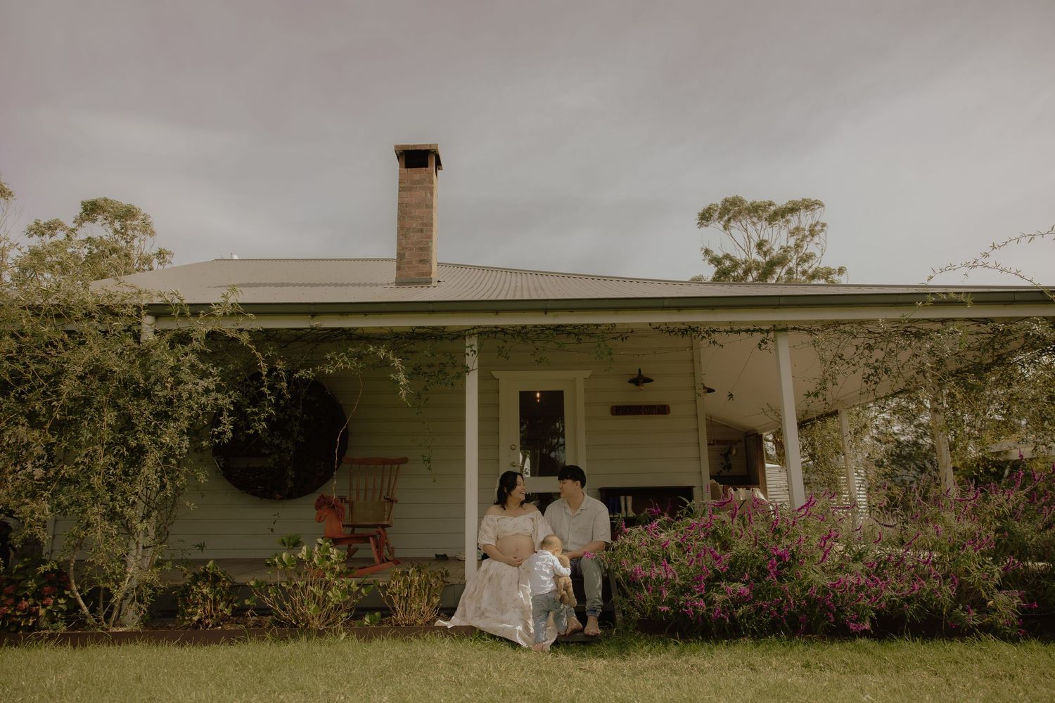 Couple sitting together on porch of charming white cottage home surrounded by lush greenery and colorful flowers.