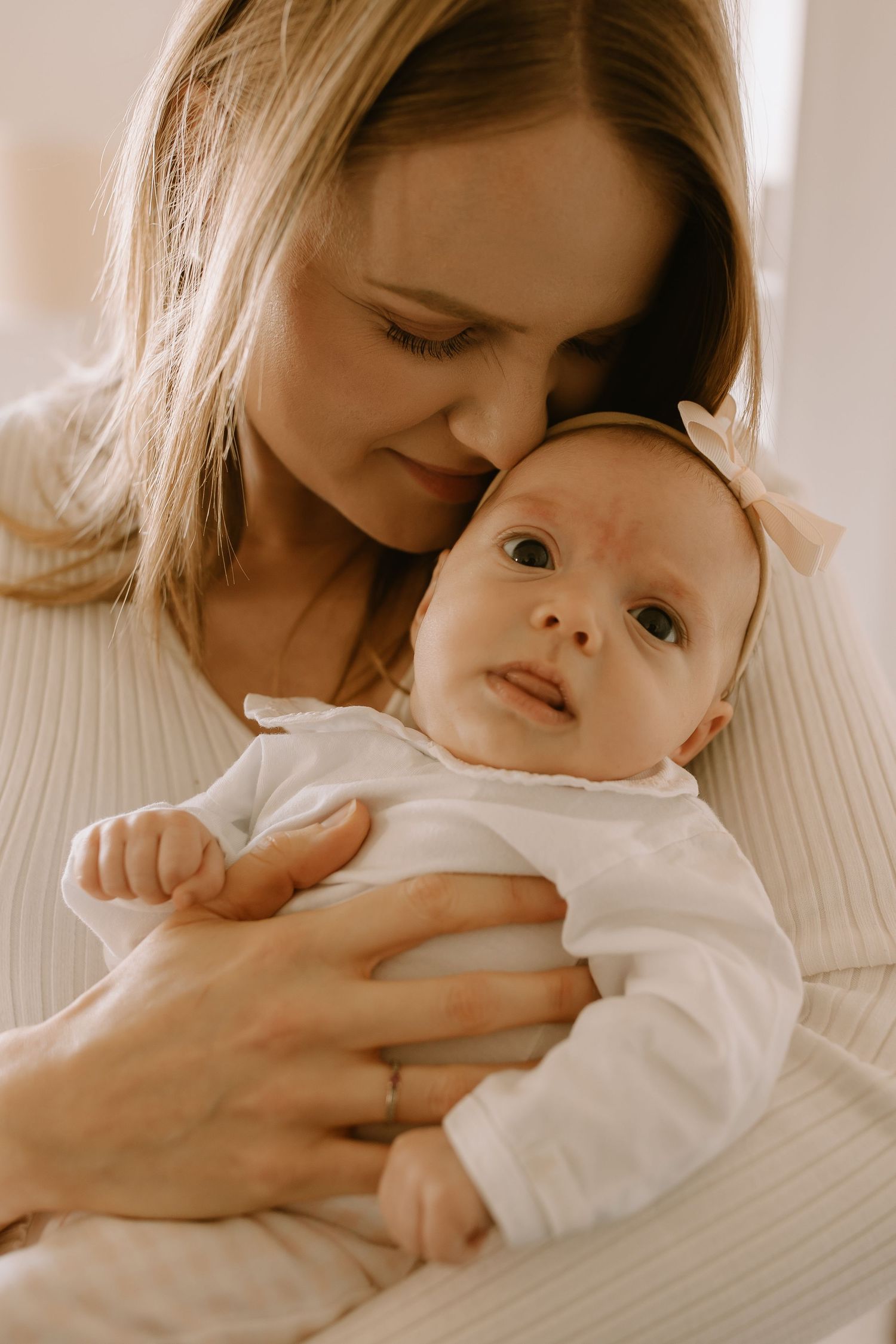 A loving mother in a white sweater embraces and gazes adoringly at her baby dressed in white.