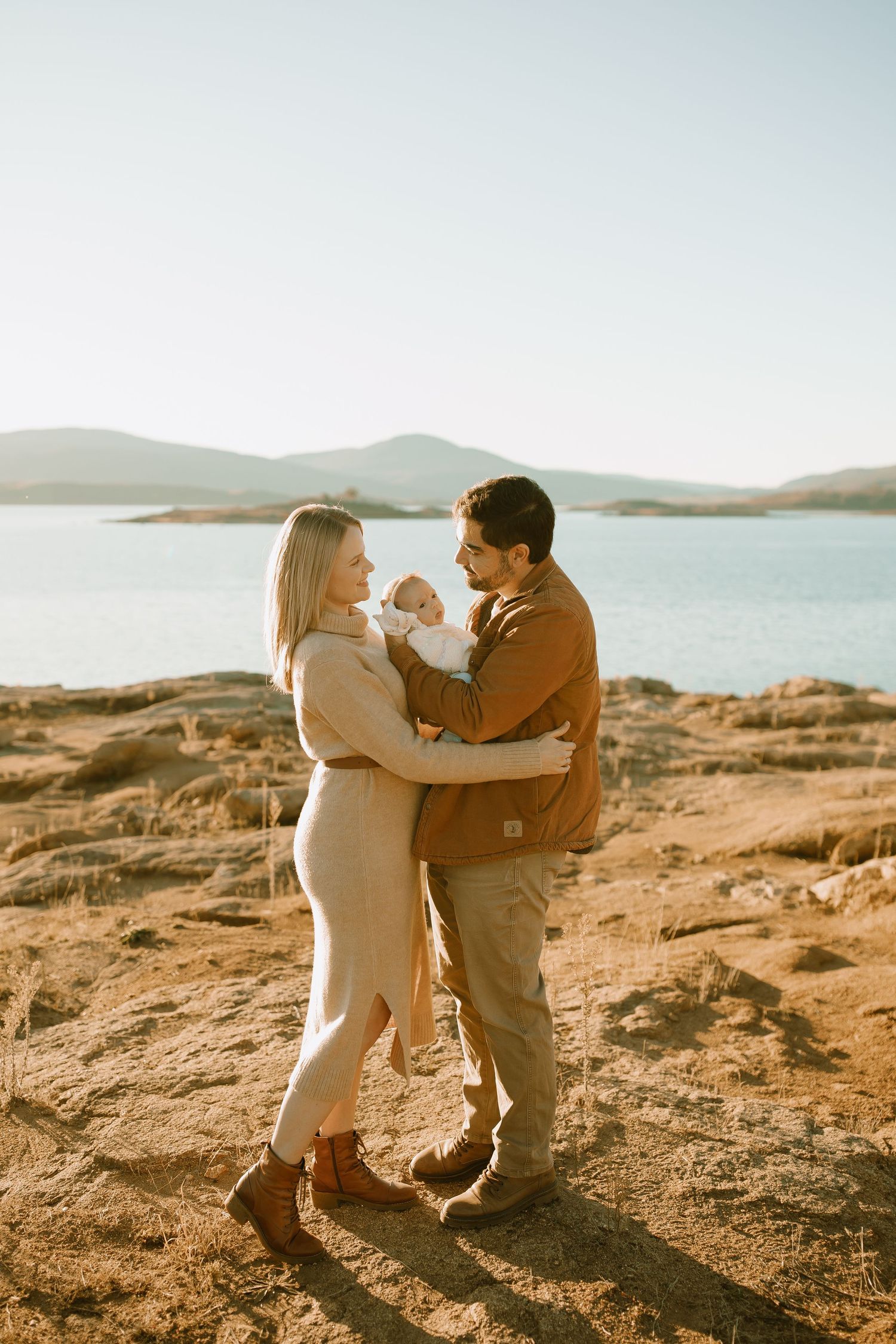 A couple embraces while holding their baby against a scenic lake backdrop at sunset.