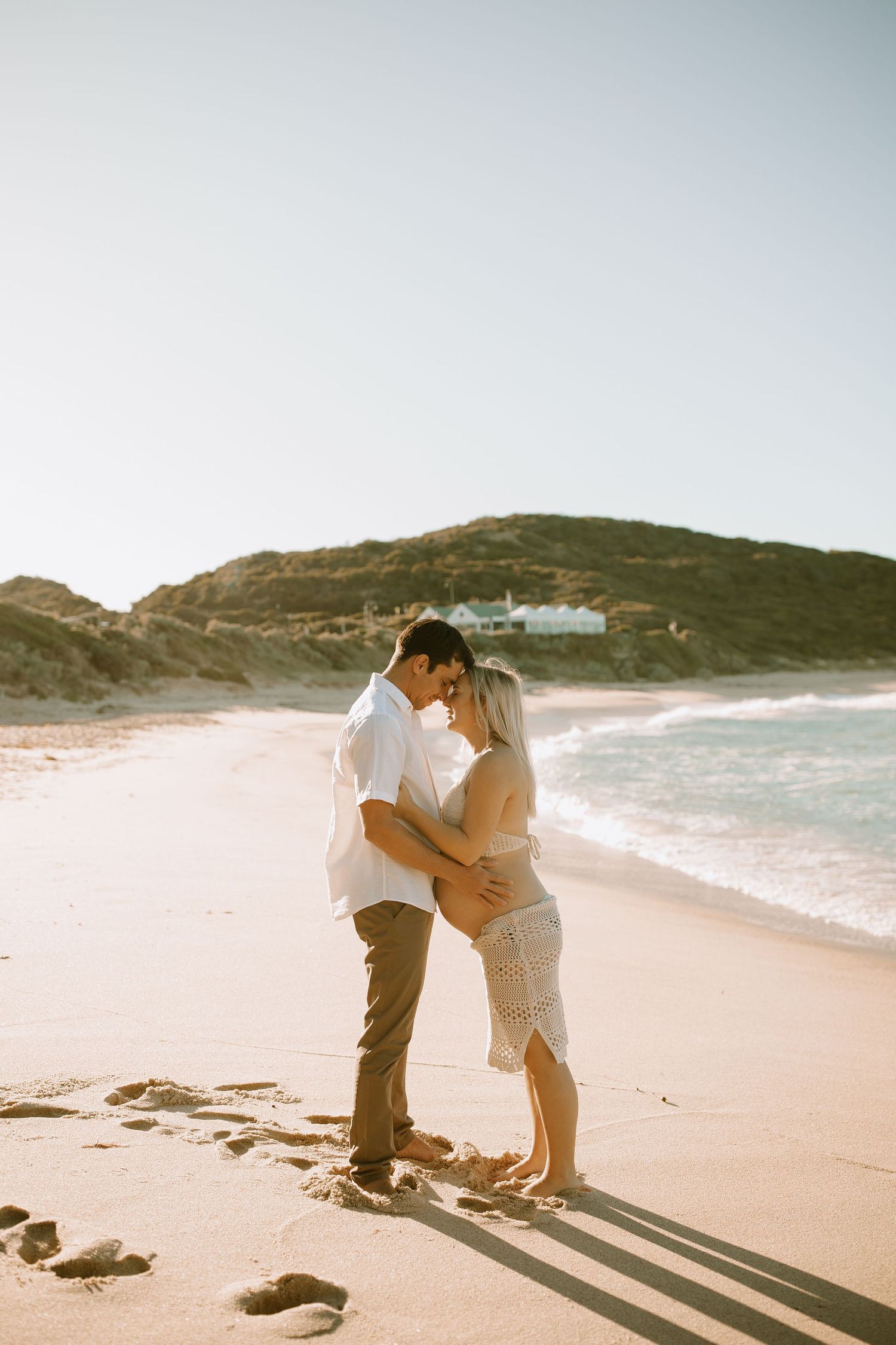 A couple embraces lovingly on a sunny beach with a hillside and ocean waves in the background.