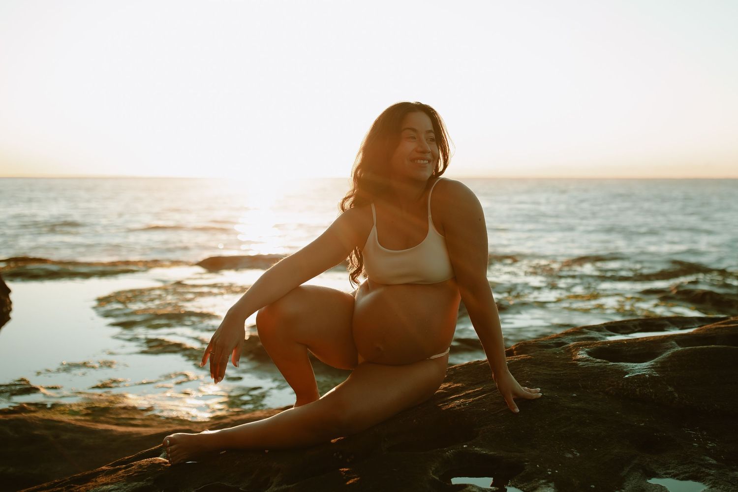A person in a white swimsuit sits on dark rocks by the ocean during golden hour sunset.
