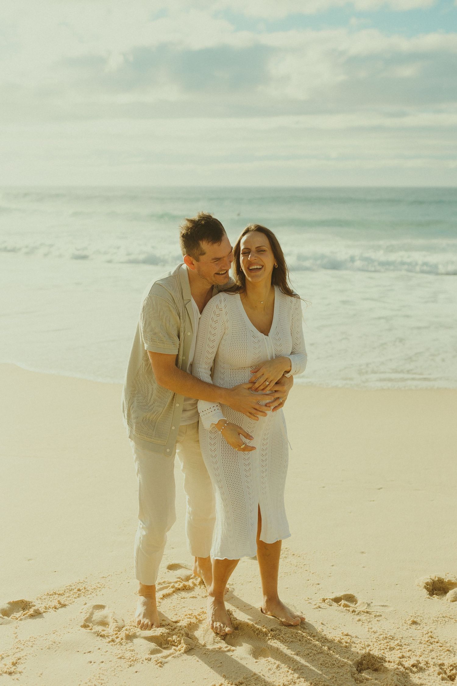 A joyful couple in casual white outfits laughing together on a sunny beach near the ocean waves.