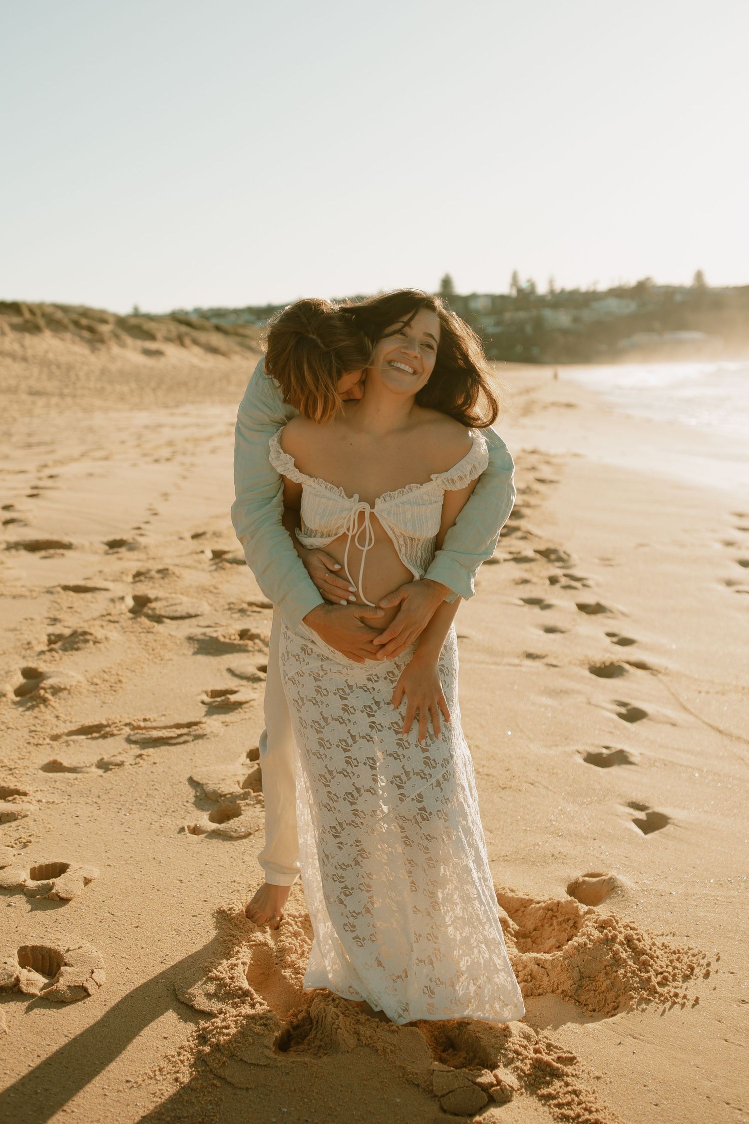 A romantic couple embraces on a sunny beach at golden hour with footprints in the sand behind them.