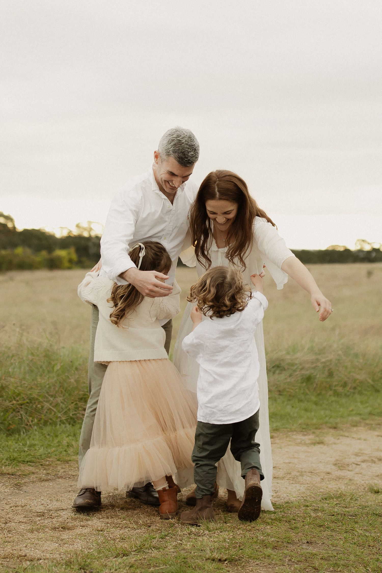 A family embraces and laughs together on a dirt path through a grassy field.