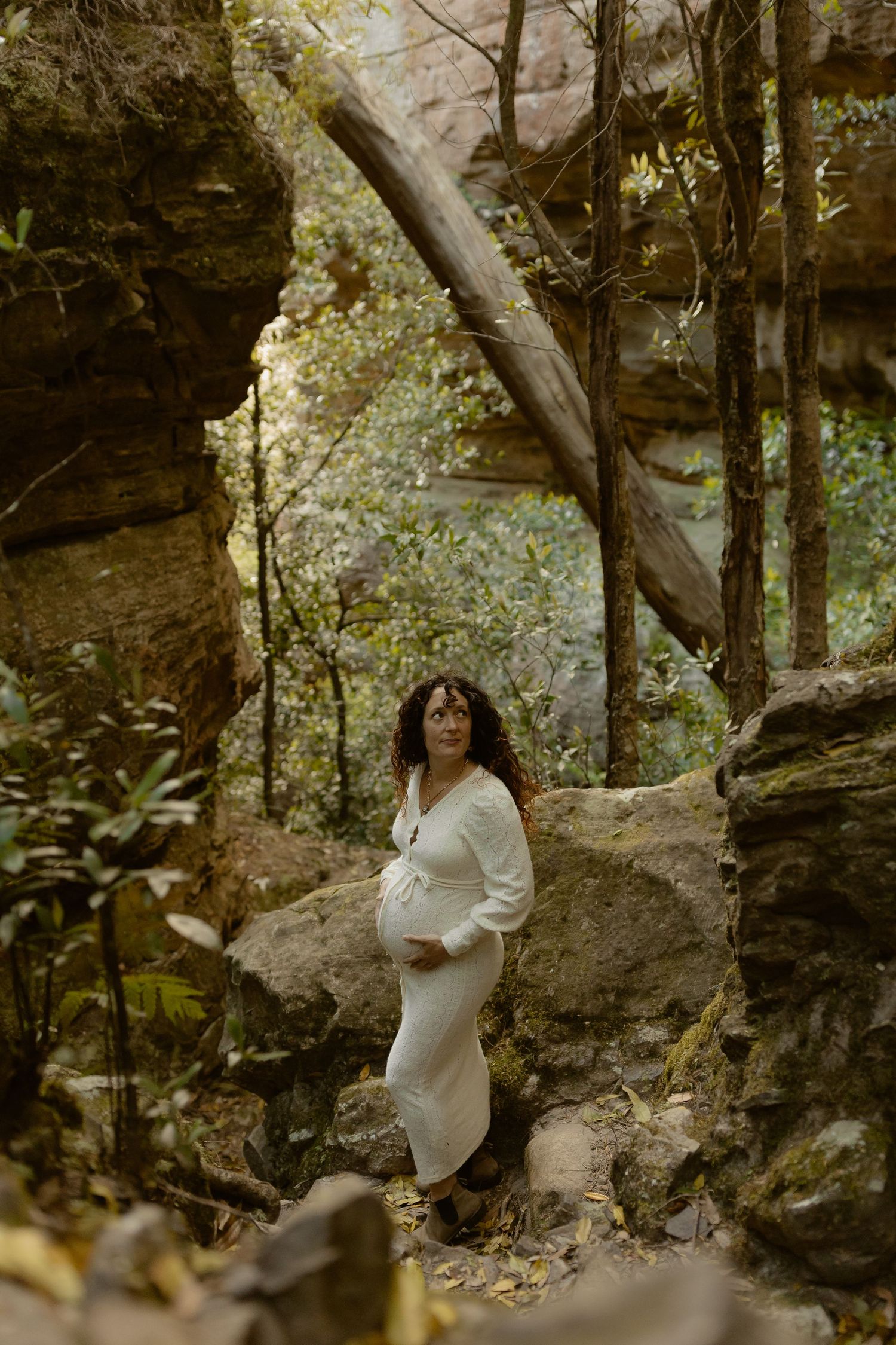 A woman in a white dress poses against dramatic rock formations in a forest setting.