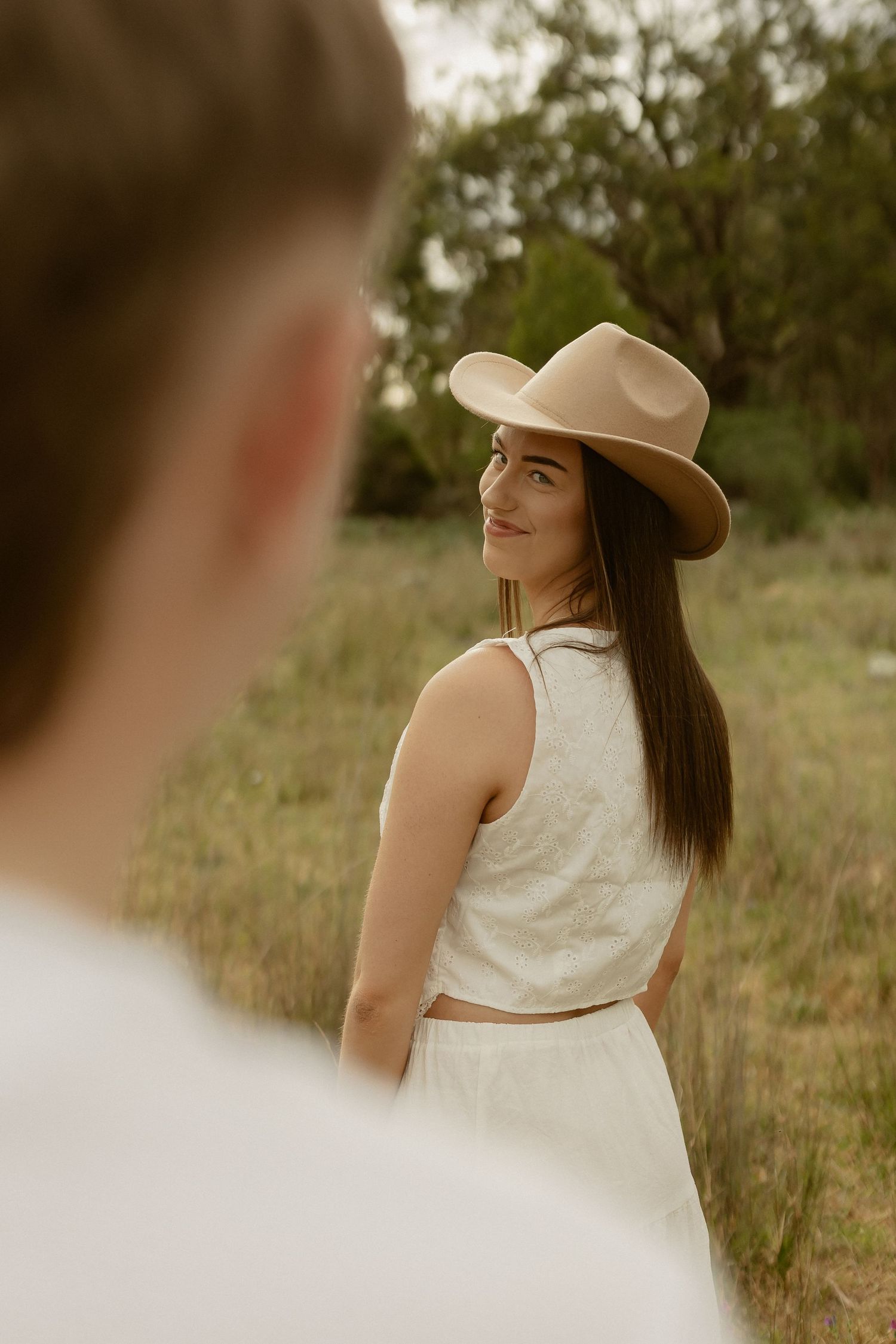 Woman in white dress and brown hat poses against natural outdoor backdrop in romantic portrait series.