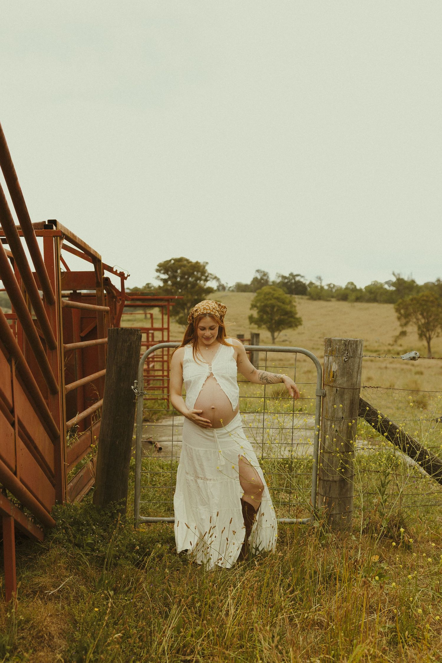 A rustic maternity photo by a farm gate featuring a white lace dress and countryside backdrop.