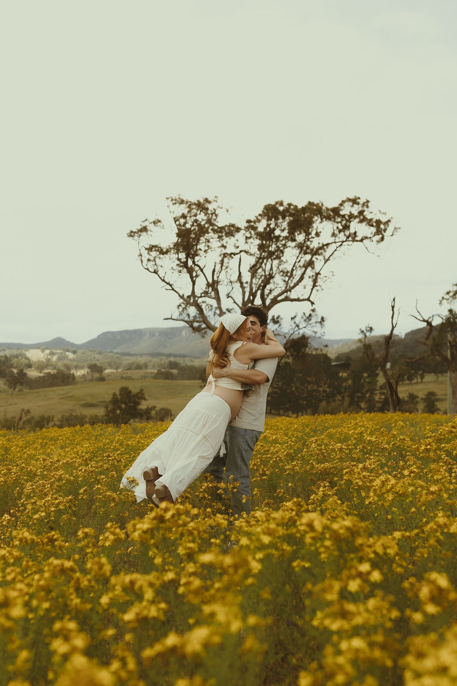 A couple embraces dramatically in a meadow of yellow flowers with a gnarled tree silhouetted against mountains.