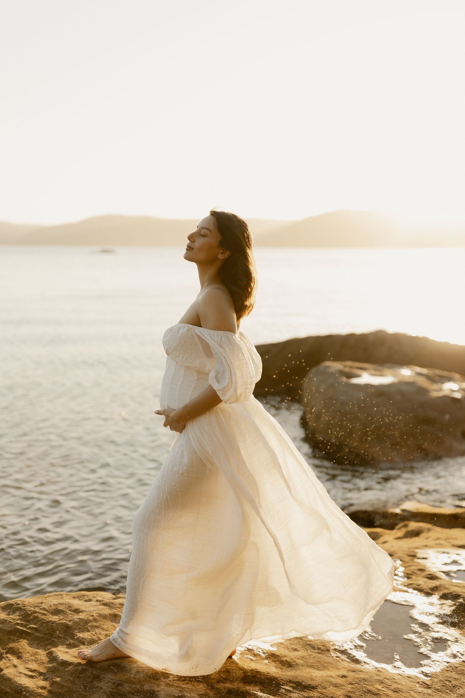 A flowing white wedding dress twirls gracefully against a sunset seascape backdrop on rocky coastal cliffs.