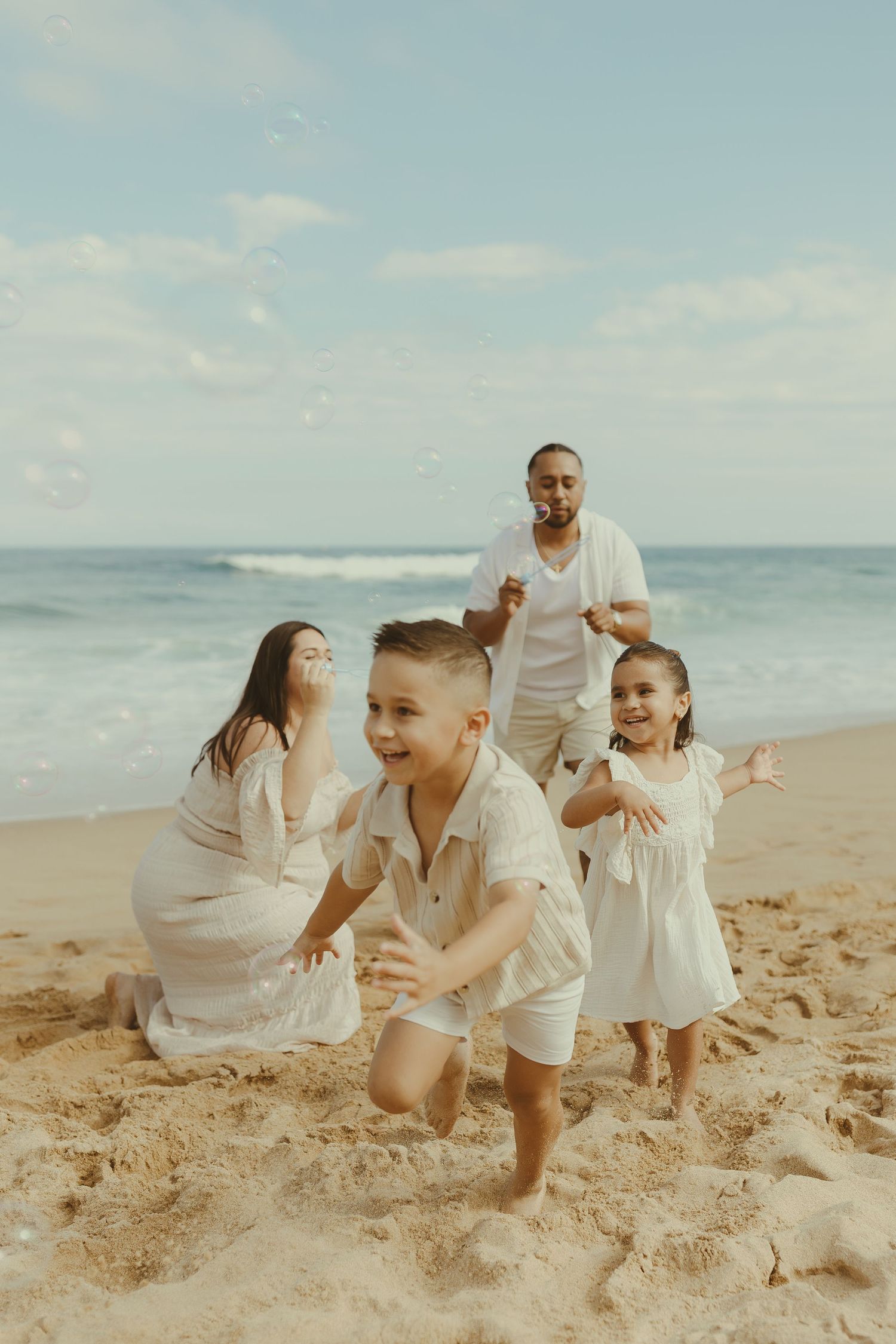 A family in white clothing plays joyfully together on a sunny beach with ocean waves in the background.