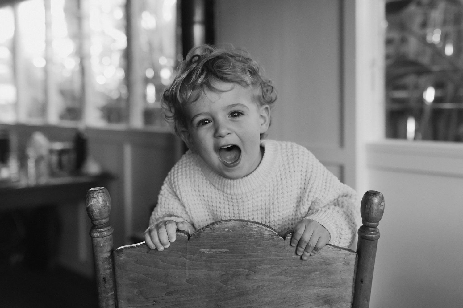 Black and white portrait of laughing toddler in knit sweater gripping wooden chair.