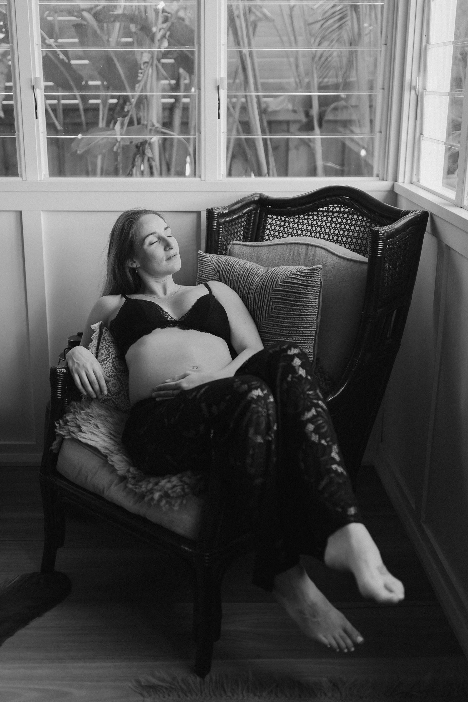 Black and white maternity photo showing pregnant woman reclining on antique bench in sun-filled room.