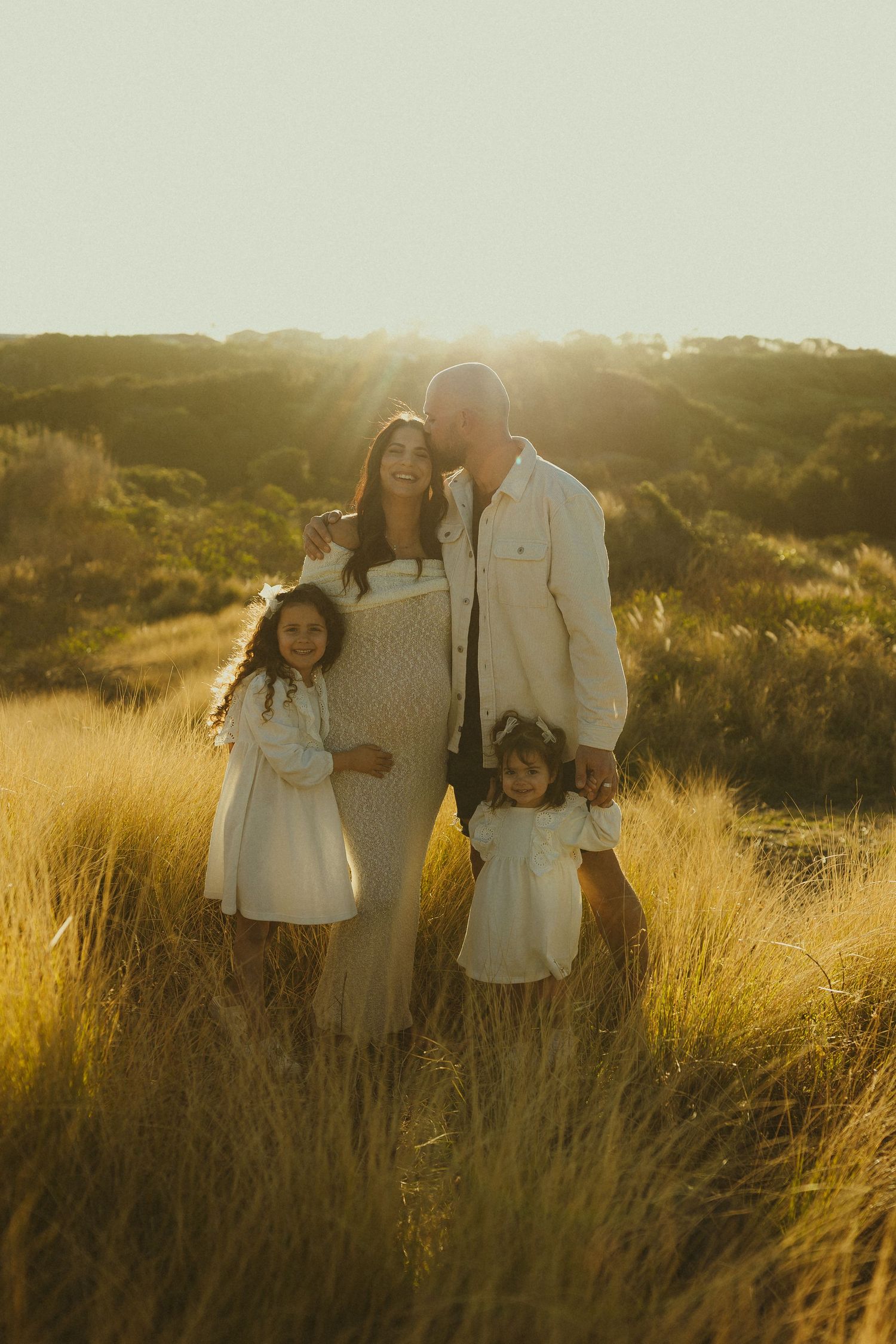 A family of four poses together in a golden field at sunset with warm backlighting creating a dreamy atmosphere.